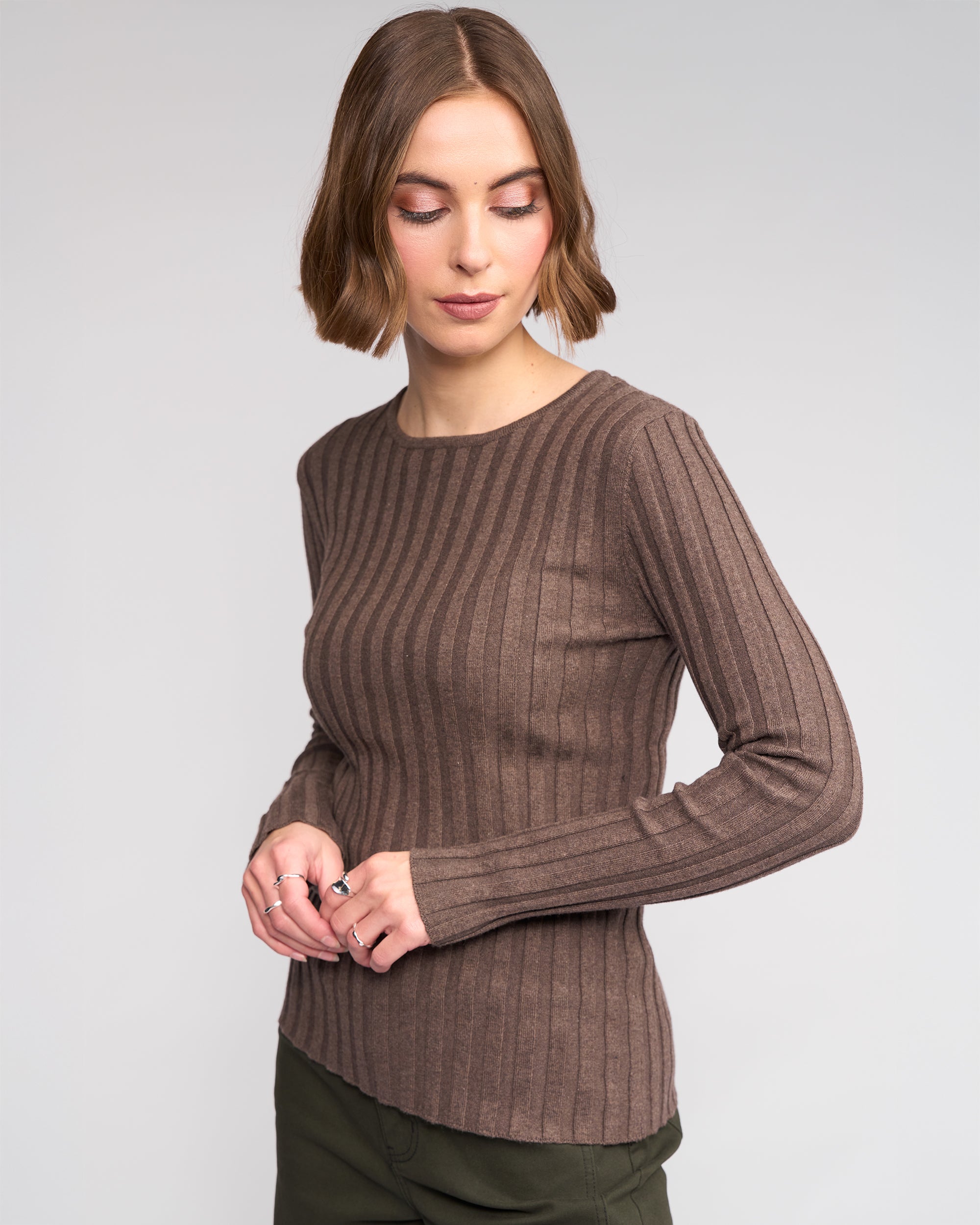 A woman with short brown hair wears Knewe Label’s Felix Sweater in Truffle and olive green pants, standing against a light gray background while looking down and slightly to the side.