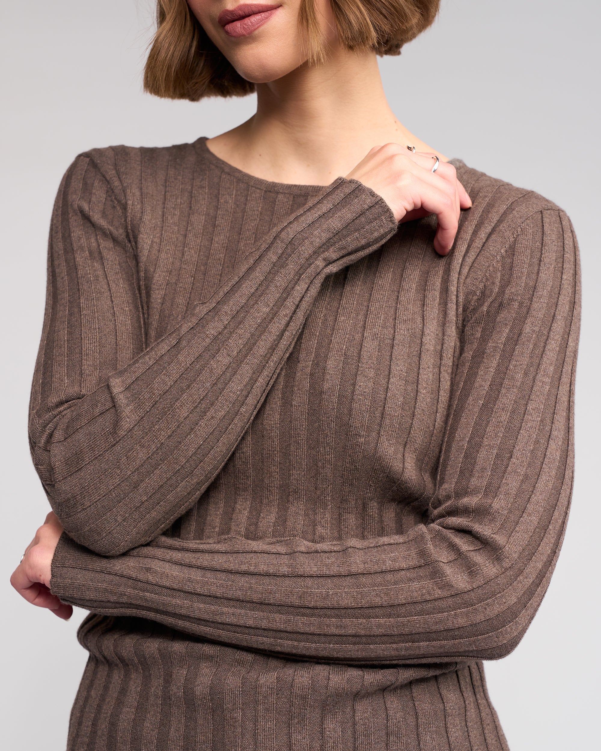 A woman with a short bob wears the Felix Sweater - Truffle by Knewe Label, a long sleeve ribbed knit, standing with arms crossed. Only her torso and part of her face are visible against a plain background.