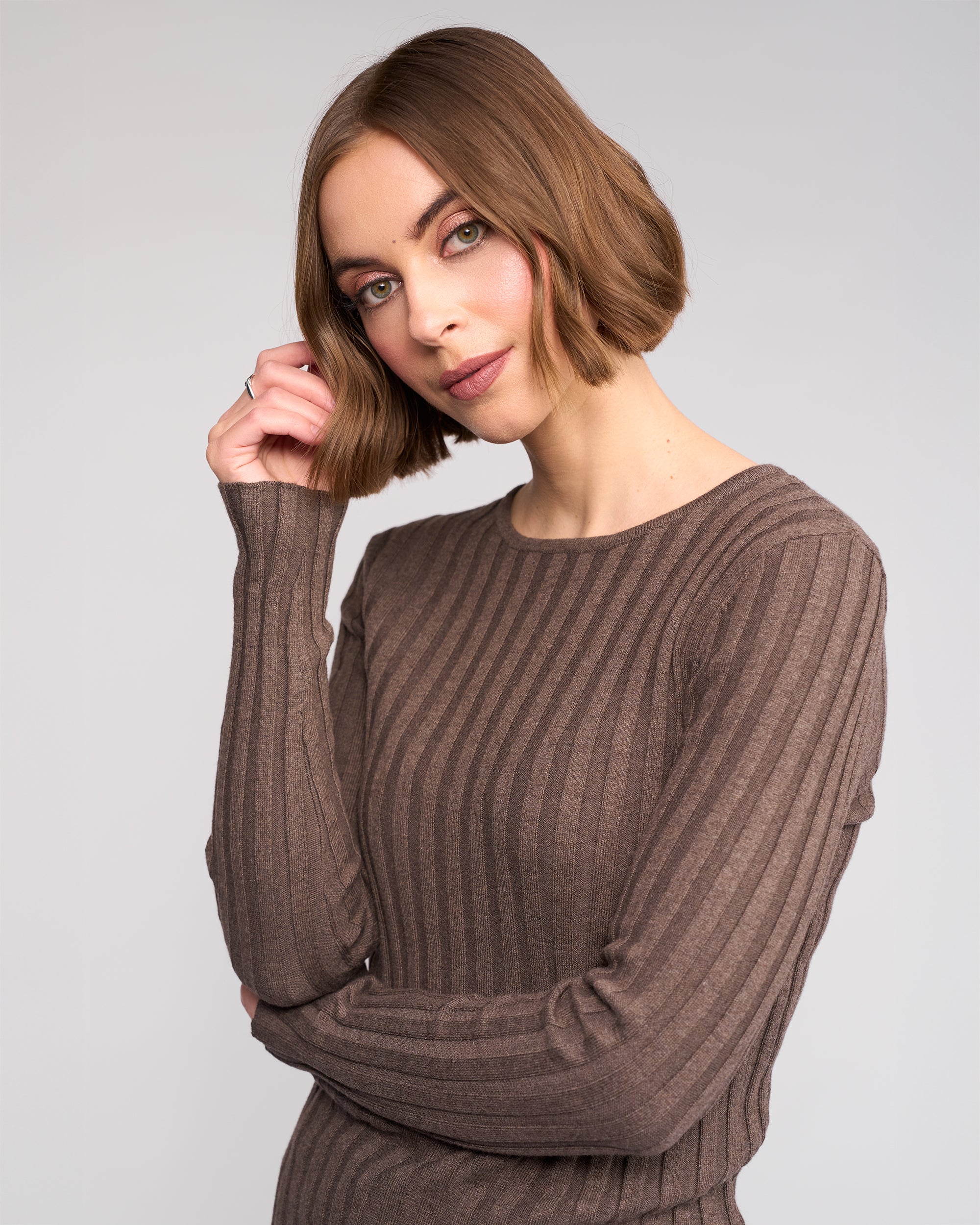 A woman with a short brown bob smiles gently, touching her hair while wearing the Knewe Label Felix Sweater in Truffle, posed against a plain light background.