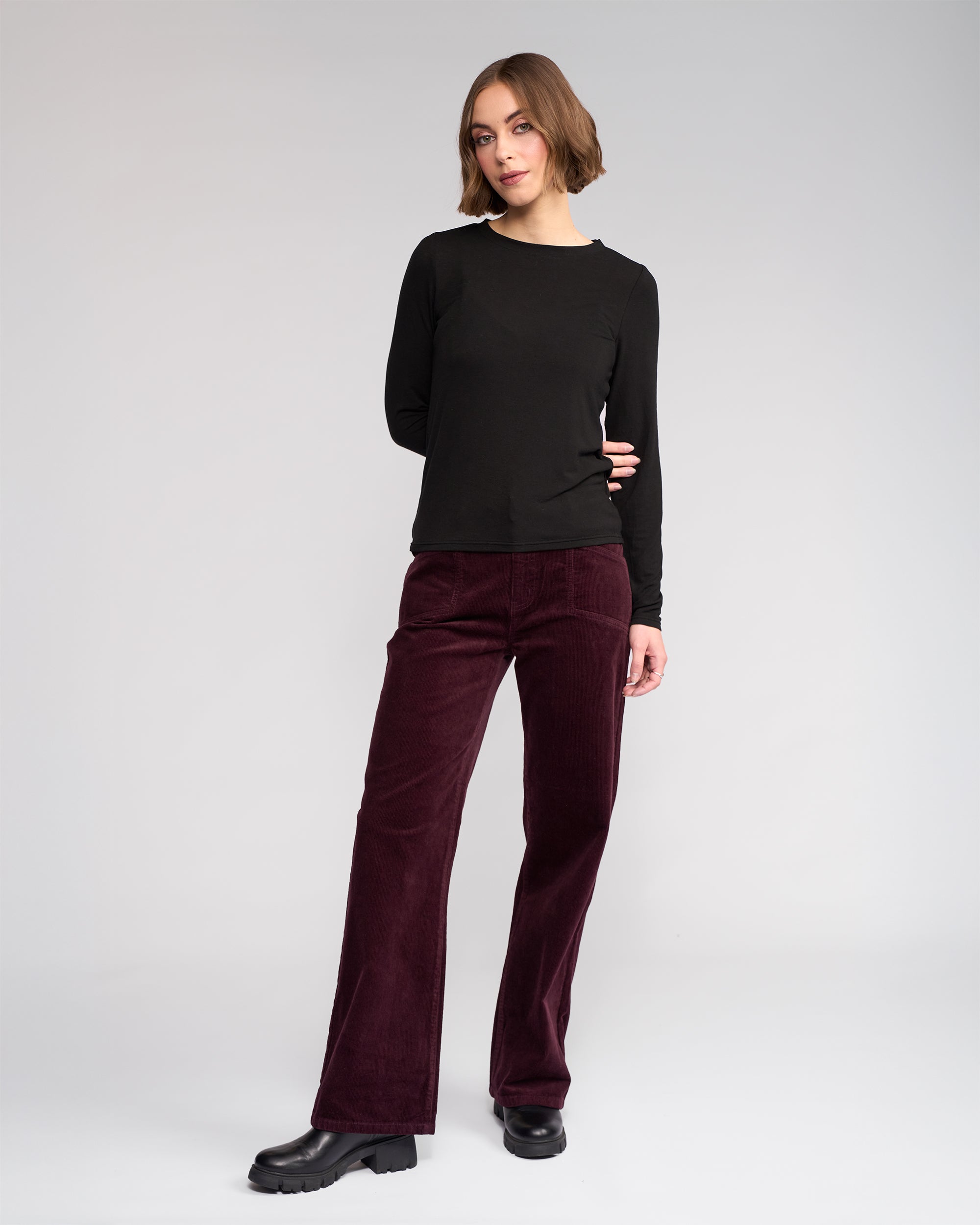 Against a plain light gray background, a person wears the Knewe Label Hero Top in black, paired with burgundy corduroy pants and black shoes. They have short brown hair, gaze slightly to the side, and keep one arm behind their back.
