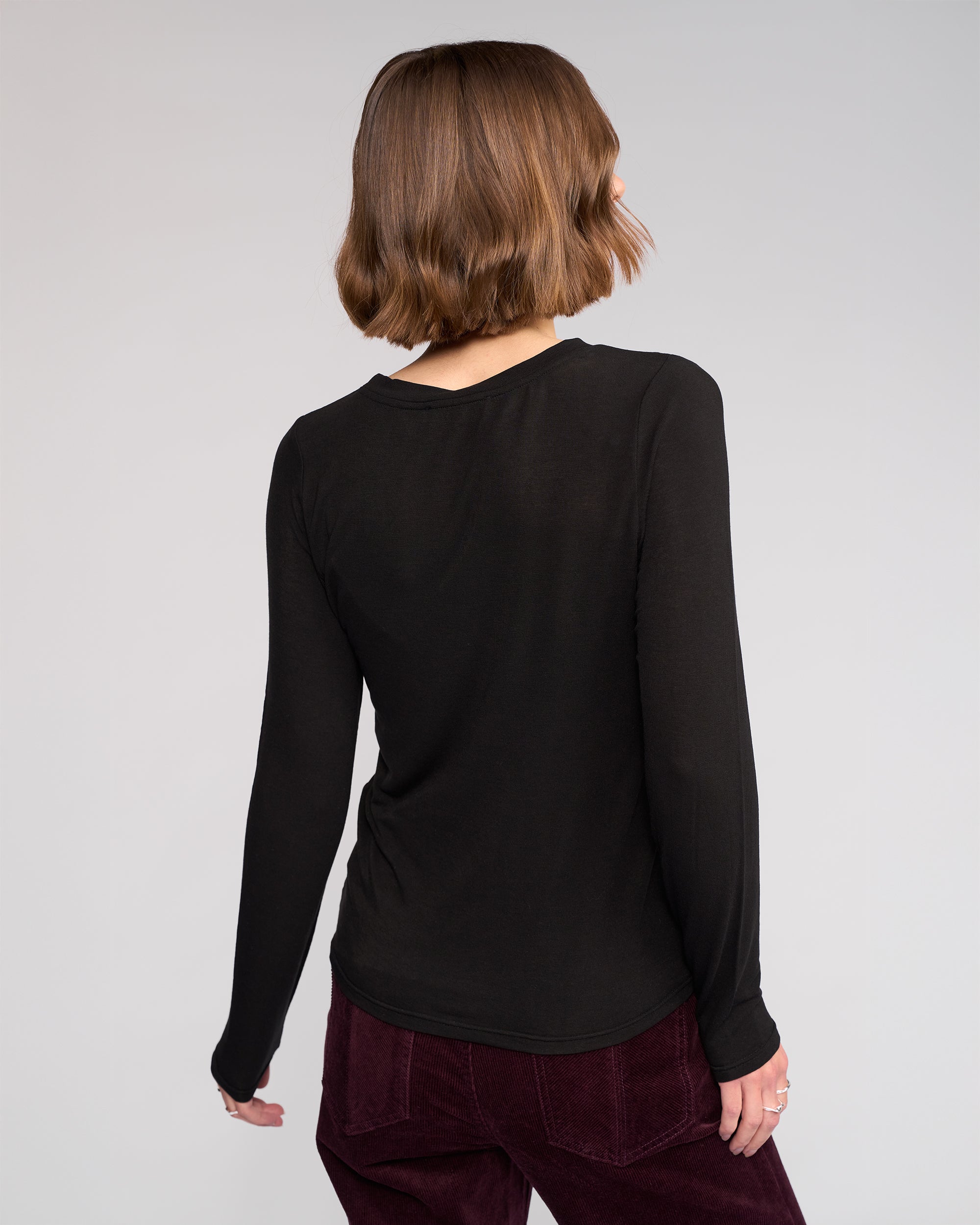 A person with short wavy brown hair stands with their back to the camera, wearing the Knewe Label Hero Top in black and burgundy pants against a plain light gray background.