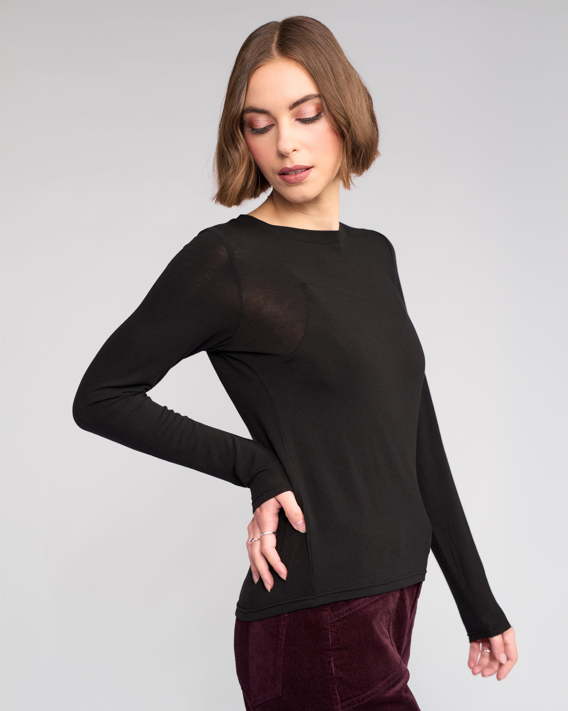 A woman with a short bob wears the Knewe Label Hero Top in black with burgundy velvet pants, standing against a plain light gray background and looking down with her hand on her hip.