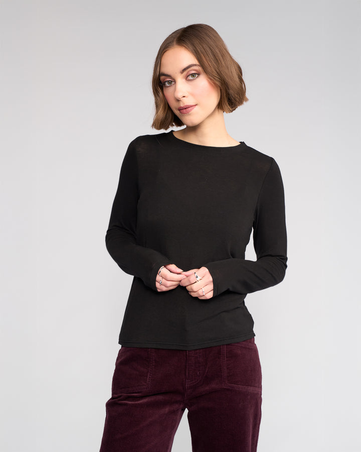 A woman with chin-length brown hair wears the Knewe Label Hero Top in black with burgundy pants, standing against a plain light gray background, hands loosely clasped.