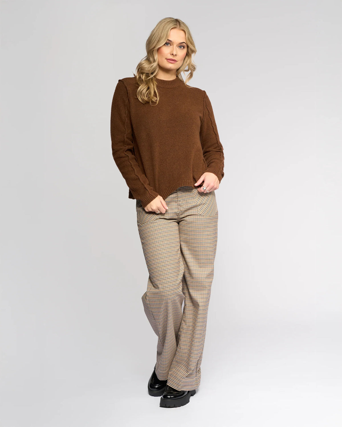 A person with long blonde hair wears the Knewe Label North Sweater in Cedar, styled with beige plaid wide-leg pants, standing against a plain light background and looking at the camera with a relaxed expression.
