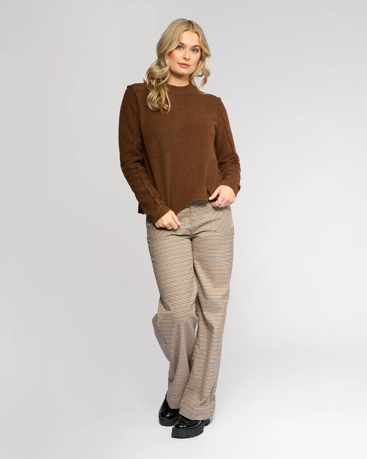 A person with long blonde hair wears the Knewe Label North Sweater in Cedar, styled with beige plaid wide-leg pants, standing against a plain light background and looking at the camera with a relaxed expression.