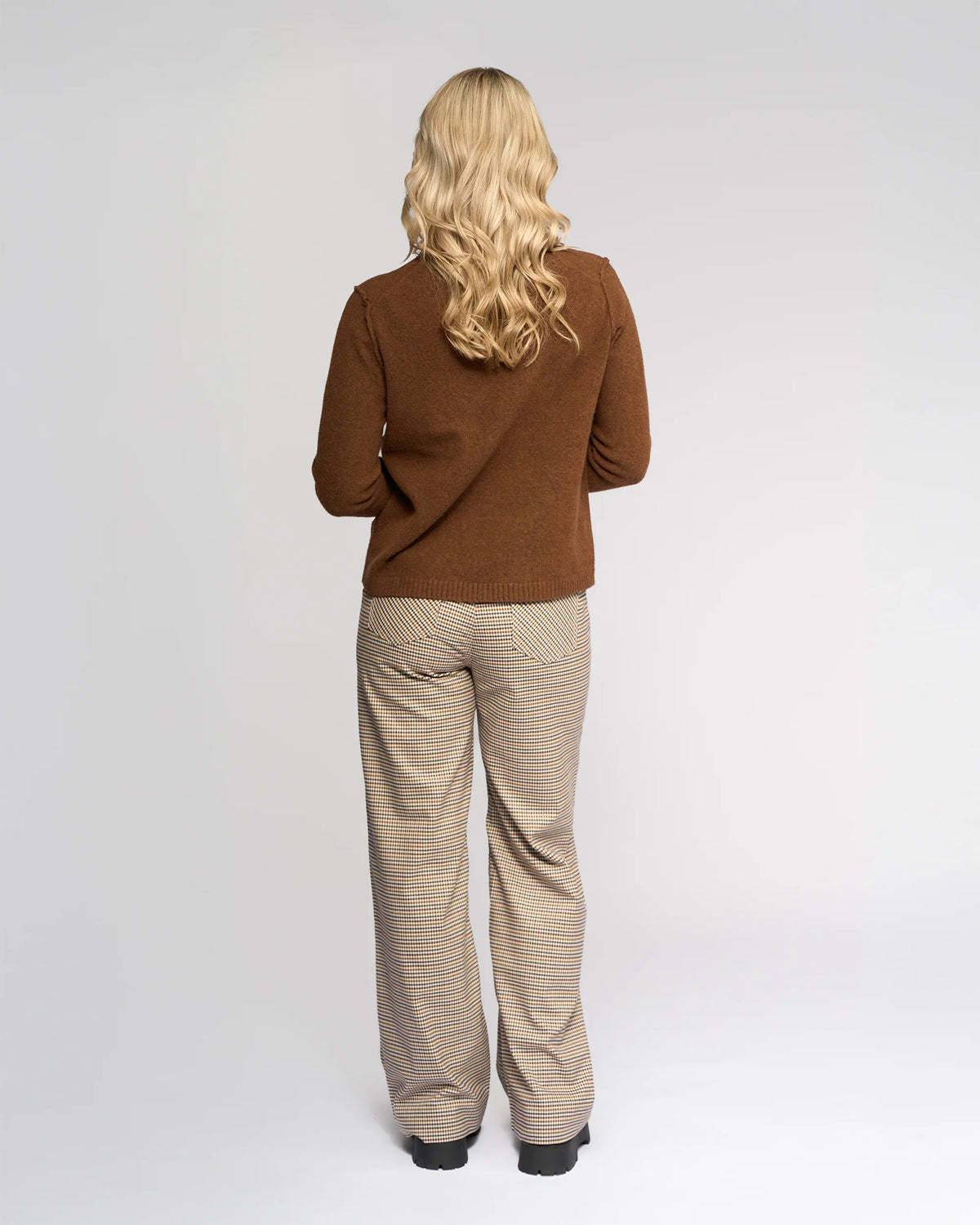 A person with long blonde hair faces away, wearing the Knewe Label North Sweater in Cedar, paired with beige checkered pants and black shoes, against a plain light gray background.