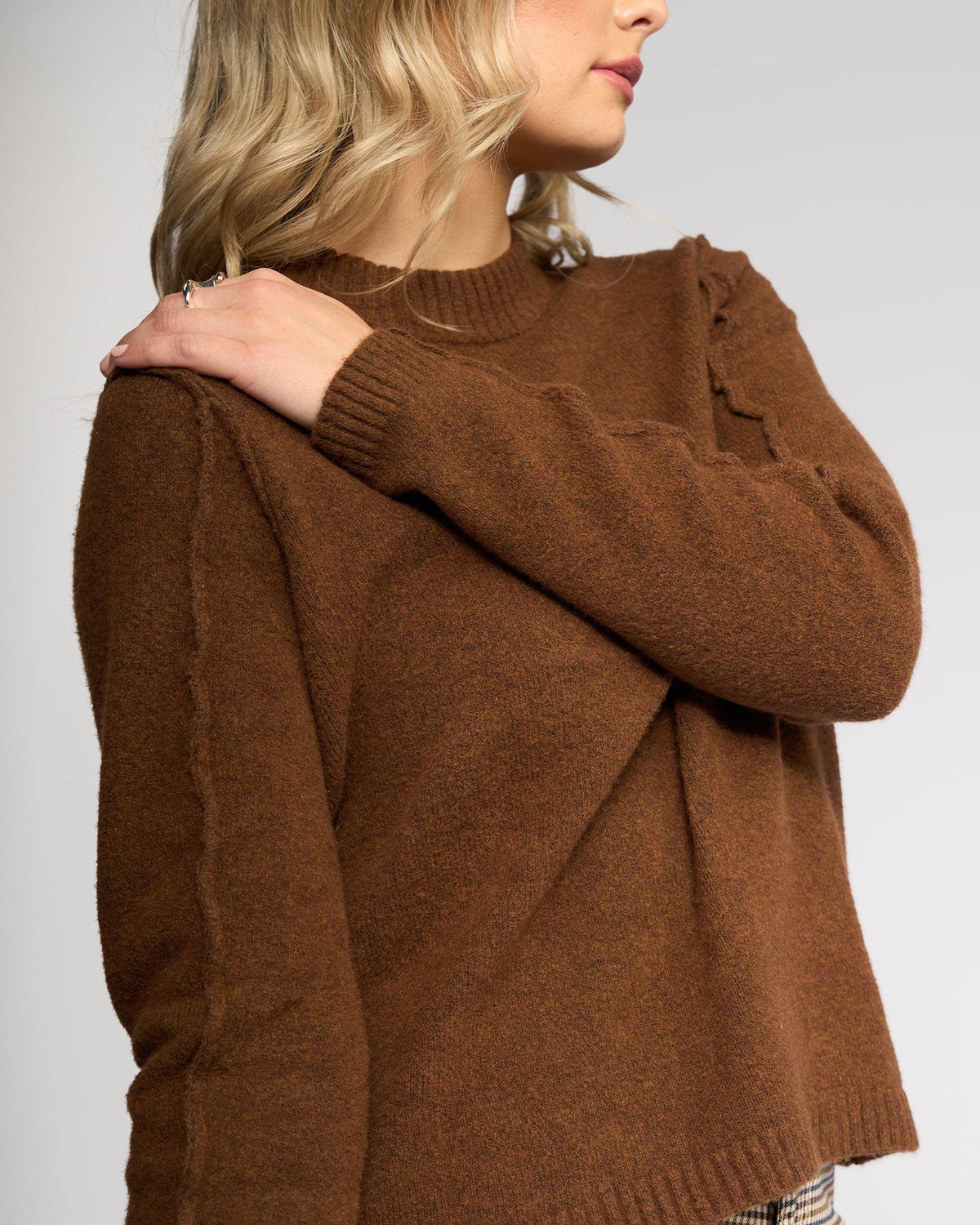 A woman with wavy blonde hair wears the North Sweater in Cedar by Knewe Label, resting her left hand on her right shoulder. Her face is partially turned away against a plain, light background.