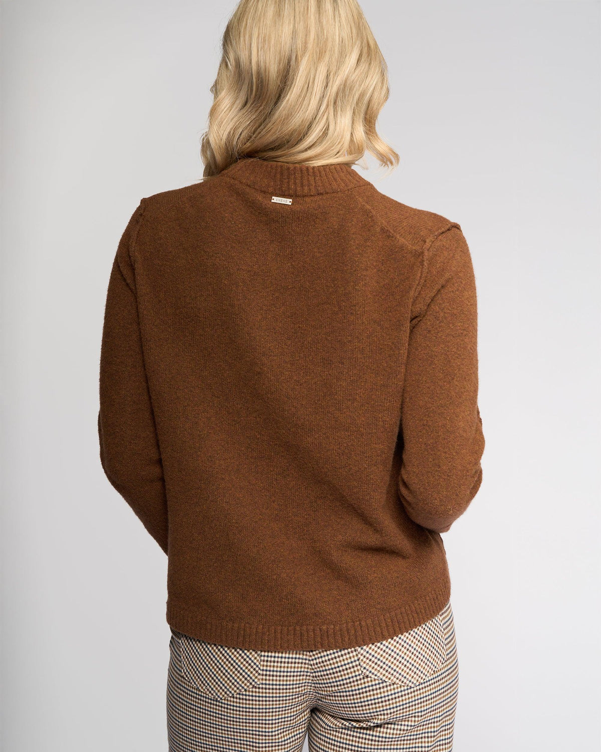 A person with blonde hair, seen from behind, wears the Knewe Label North Sweater in Cedar and checkered pants against a plain light-colored background.