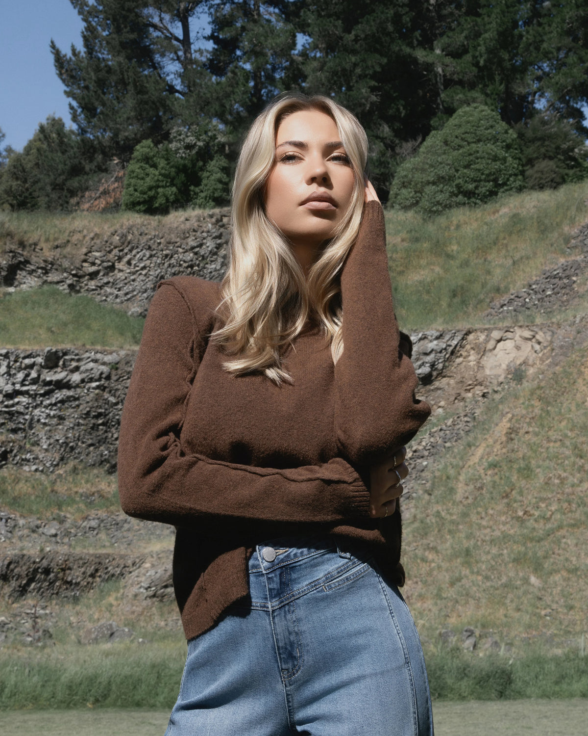 A woman with long blonde hair wears the Knewe Label North Sweater in Cedar and blue jeans, standing outdoors by a grassy hill and trees, looking confidently at the camera with one hand touching her face.