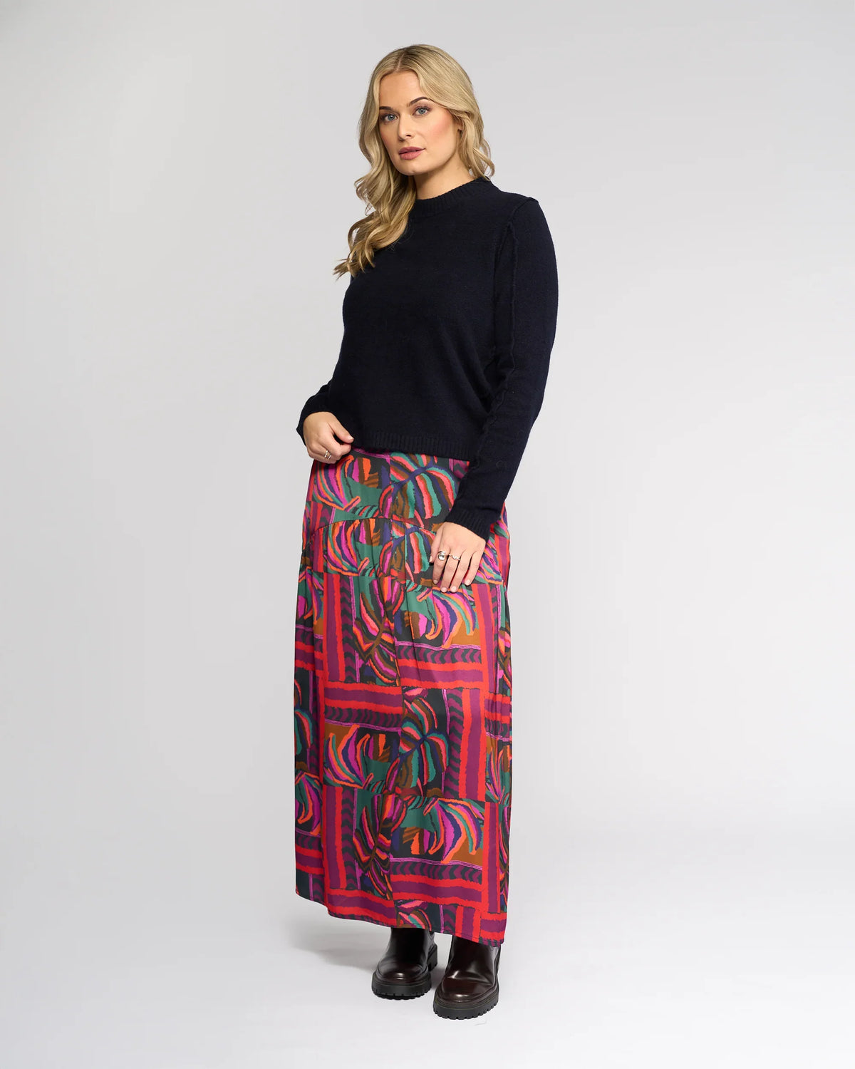 A woman with wavy blonde hair strikes a relaxed pose against a plain background, wearing the Knewe Label North Sweater in Ink, a colorful maxi skirt with red, pink, and green patterns, and black boots.