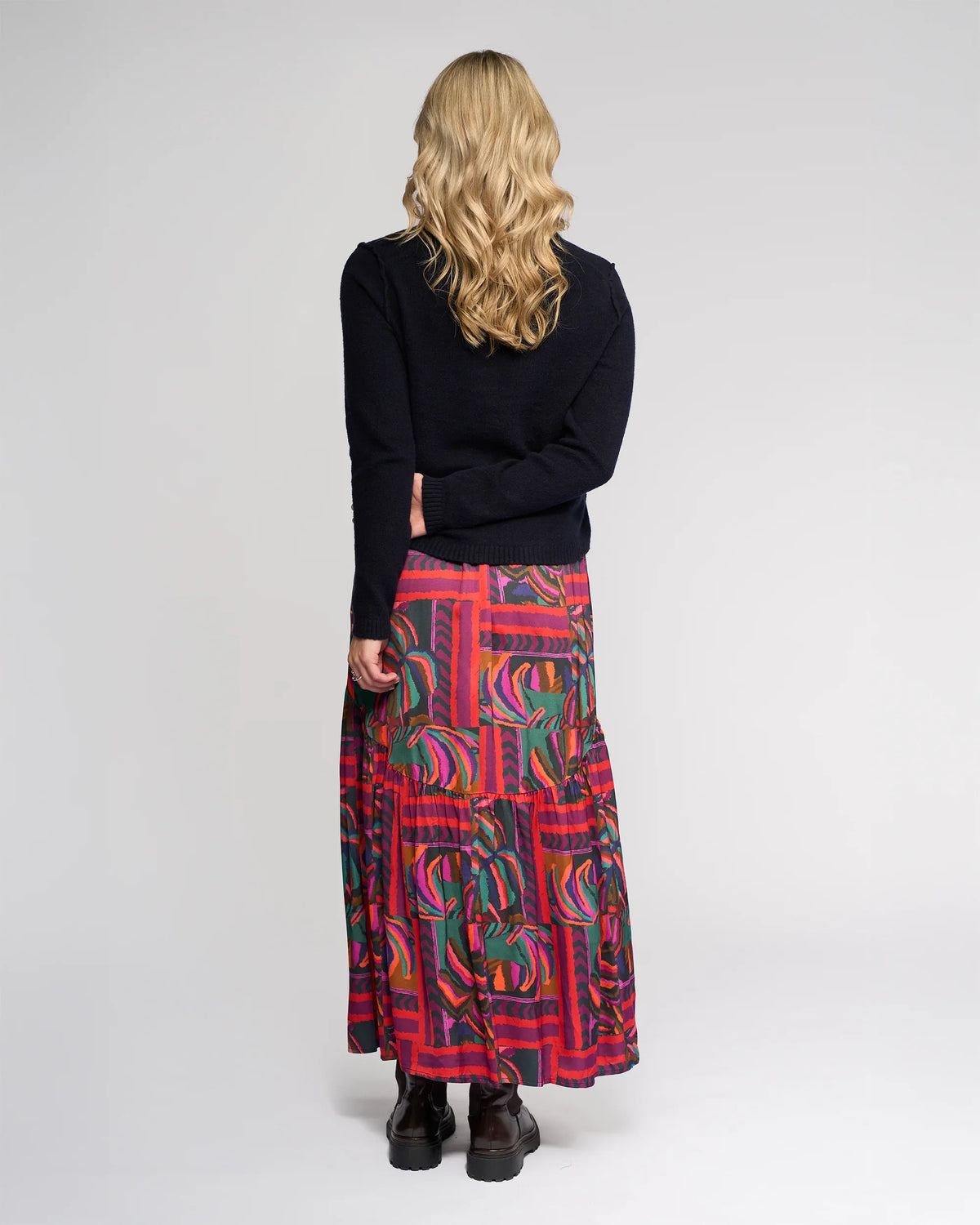 A person with wavy blonde hair wears the Knewe Label North Sweater - Ink in a relaxed fit, paired with a colorful, patterned ankle-length skirt, and stands facing away from the camera against a plain light background.