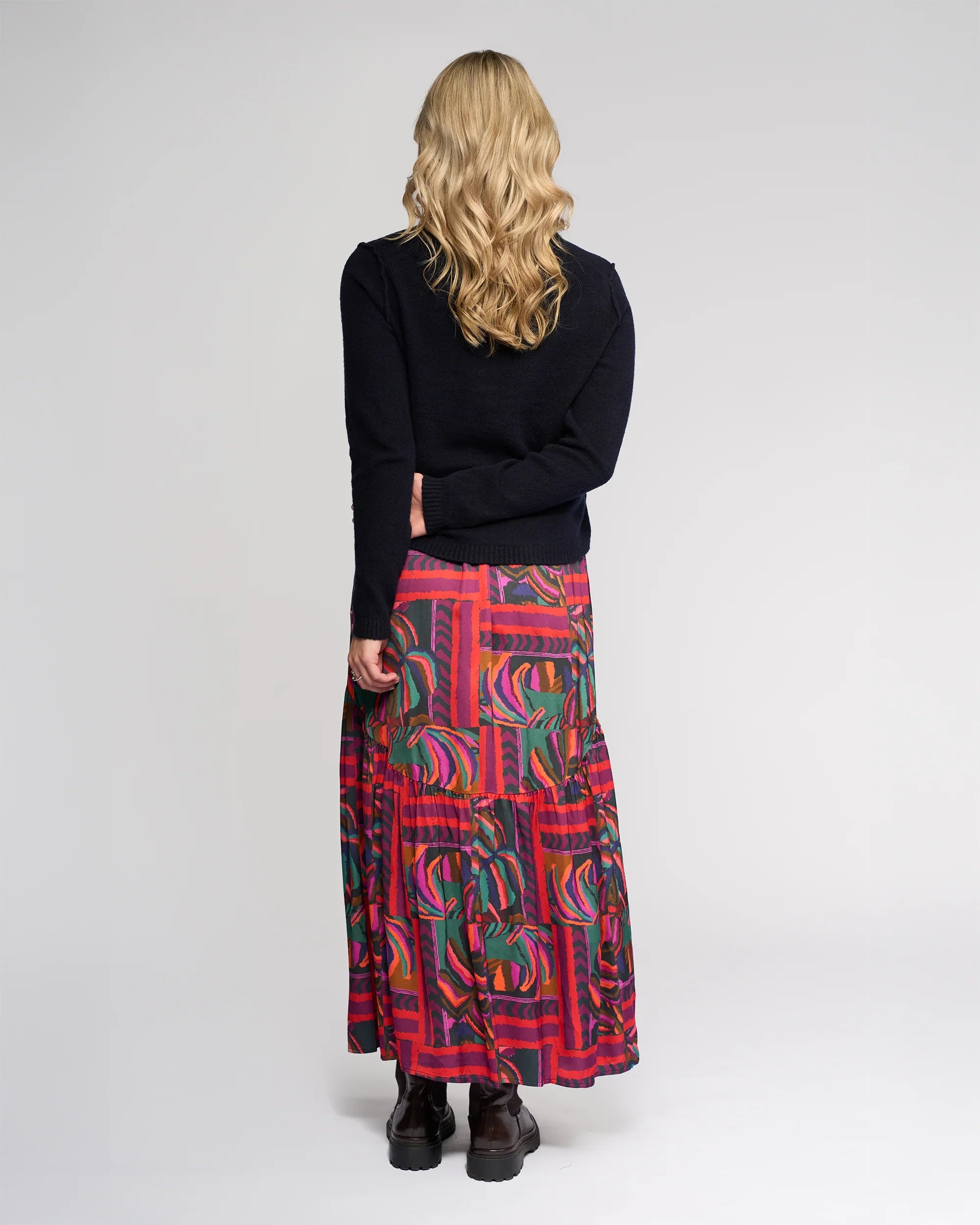 A person with wavy blonde hair wears the Knewe Label North Sweater - Ink in a relaxed fit, paired with a colorful, patterned ankle-length skirt, and stands facing away from the camera against a plain light background.