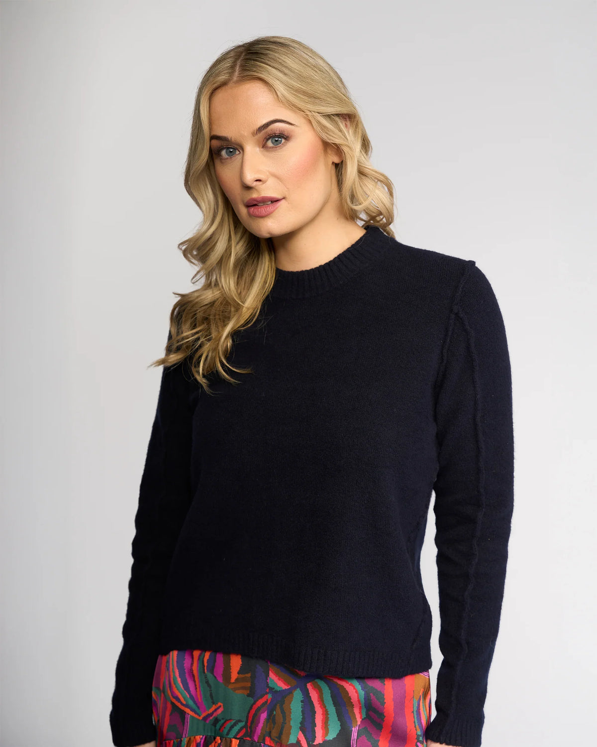 A woman with long blonde hair wears the Knewe Label North Sweater in Ink with exposed seam detailing, paired with a colorful skirt. She stands against a light gray background, looking to the side with a neutral expression.