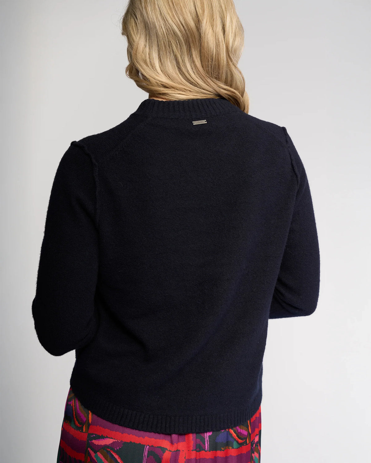 A person with long blonde hair is seen from the back, wearing the Knewe Label North Sweater in Ink and a colorful red, purple, and blue patterned skirt against a plain light background.