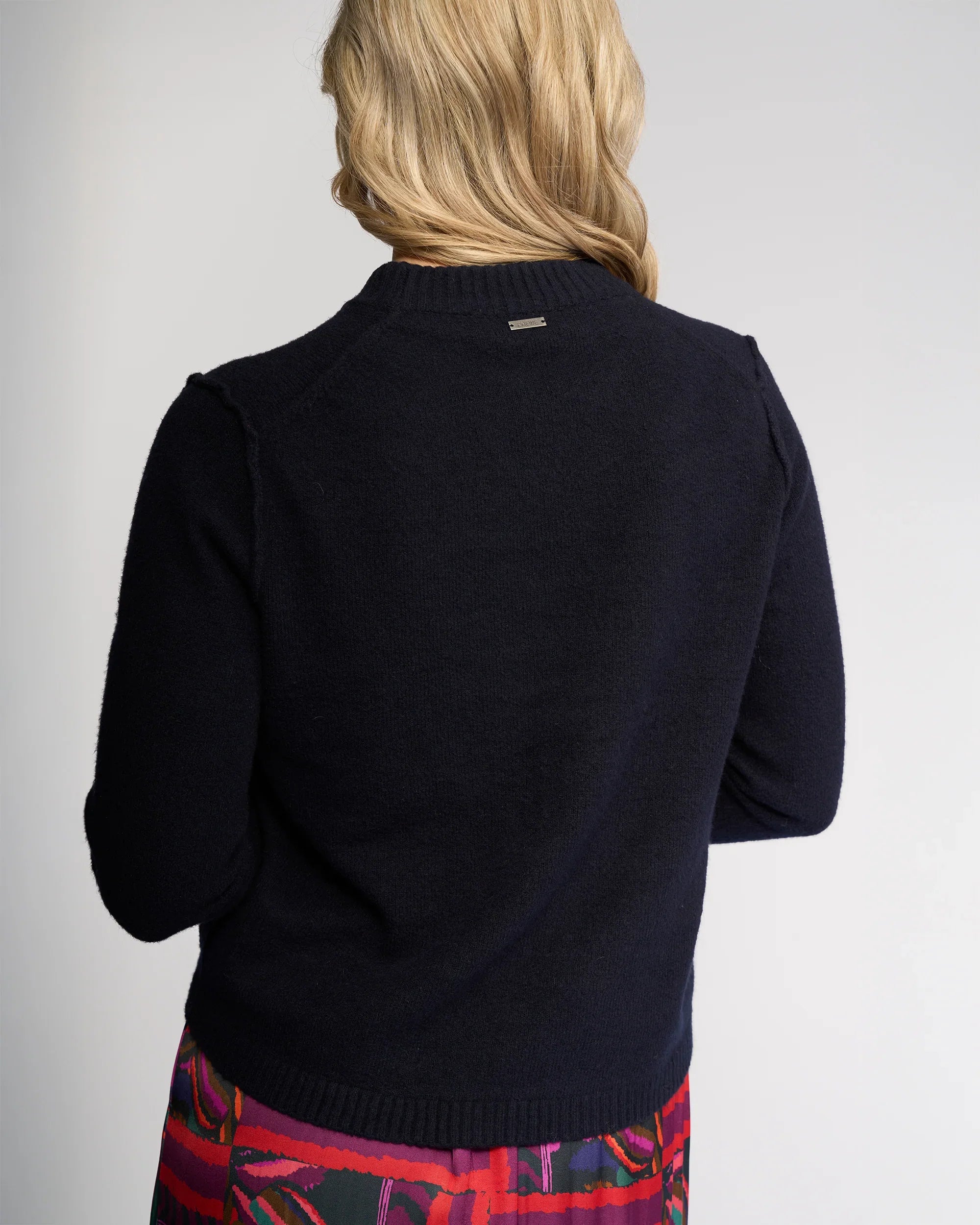A person with long blonde hair is seen from the back, wearing the Knewe Label North Sweater in Ink and a colorful red, purple, and blue patterned skirt against a plain light background.