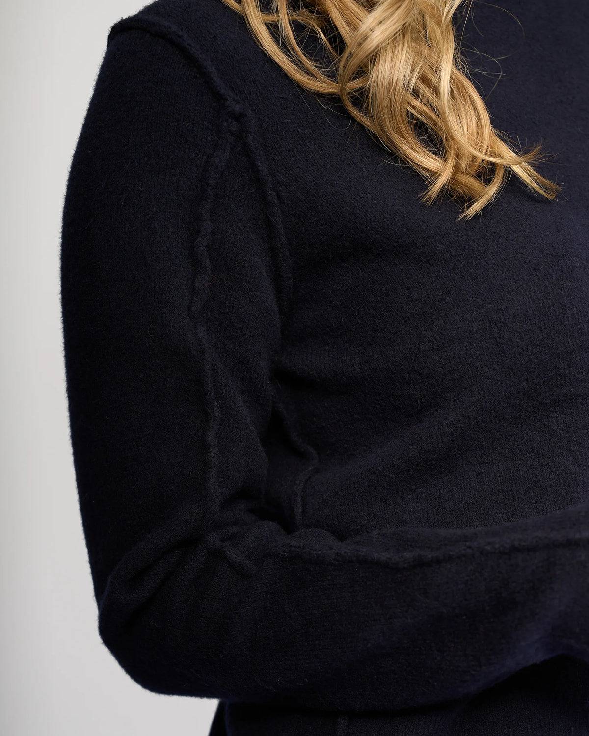 Close-up of a person wearing the Knewe Label North Sweater in Ink. Only their upper torso and part of the arm are visible; their face is not shown. The dark navy knit features exposed seam detailing, with light brown wavy hair over one shoulder.