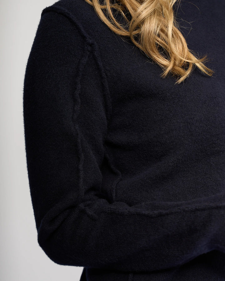 Close-up of a person wearing the Knewe Label North Sweater in Ink. Only their upper torso and part of the arm are visible; their face is not shown. The dark navy knit features exposed seam detailing, with light brown wavy hair over one shoulder.