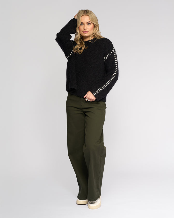 A woman with blonde hair poses against a plain background wearing the Knewe Label Lennox Sweater in Black/Winter White, featuring relaxed fit and white sleeve stitching, paired with olive wide-leg pants and cream platform shoes. One hand rests on her head.