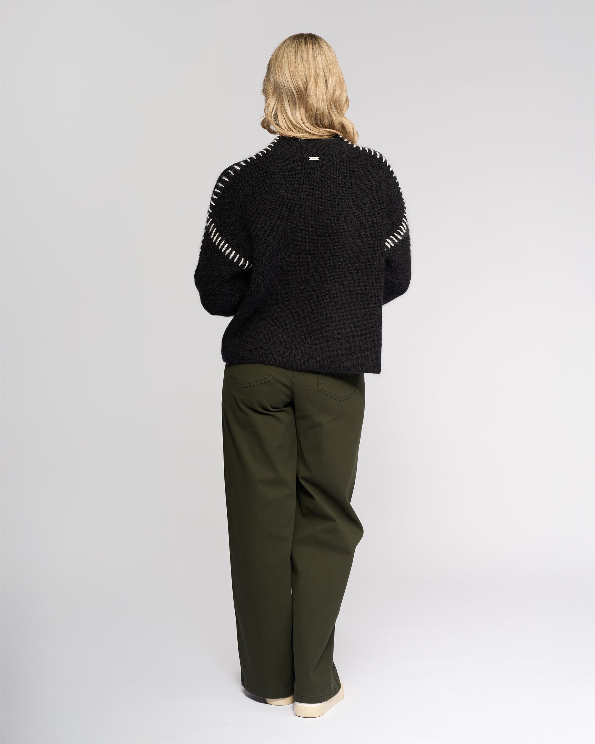 A person with blonde hair faces away, wearing the Knewe Label Lennox Sweater in Black/Winter White, paired with olive green wide-leg pants and cream shoes against a plain light background.