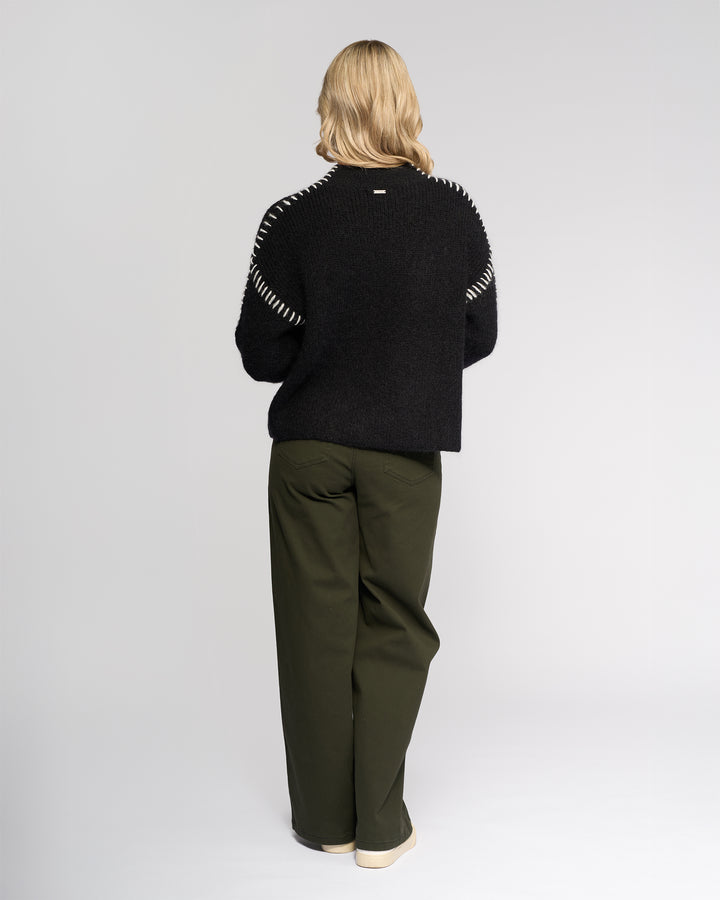 A person with blonde hair faces away, wearing the Knewe Label Lennox Sweater in Black/Winter White, paired with olive green wide-leg pants and cream shoes against a plain light background.