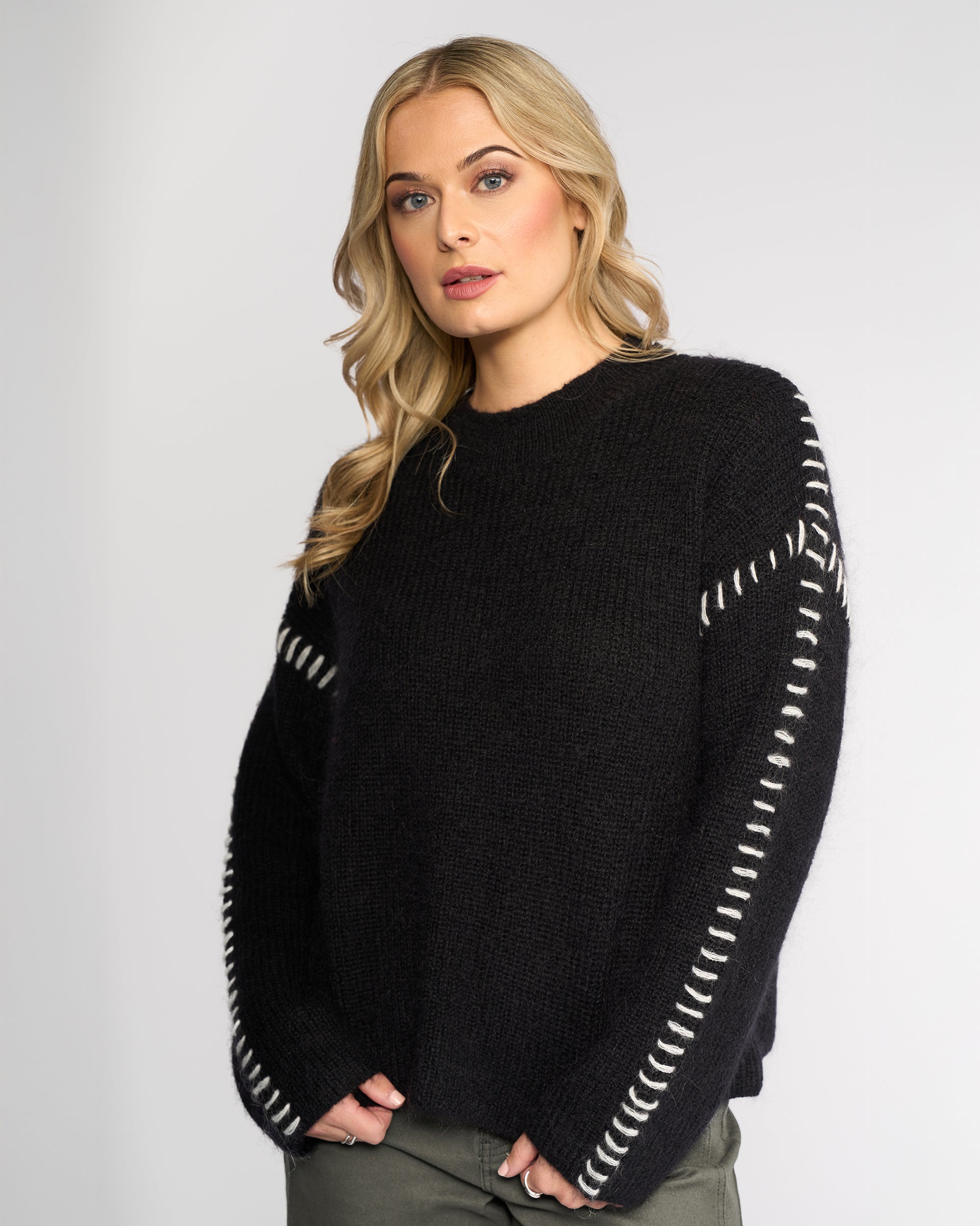 A woman with long blonde hair wears the Knewe Label Lennox Sweater in Black/Winter White, featuring white sleeve stitching, paired with dark green pants against a plain light background.