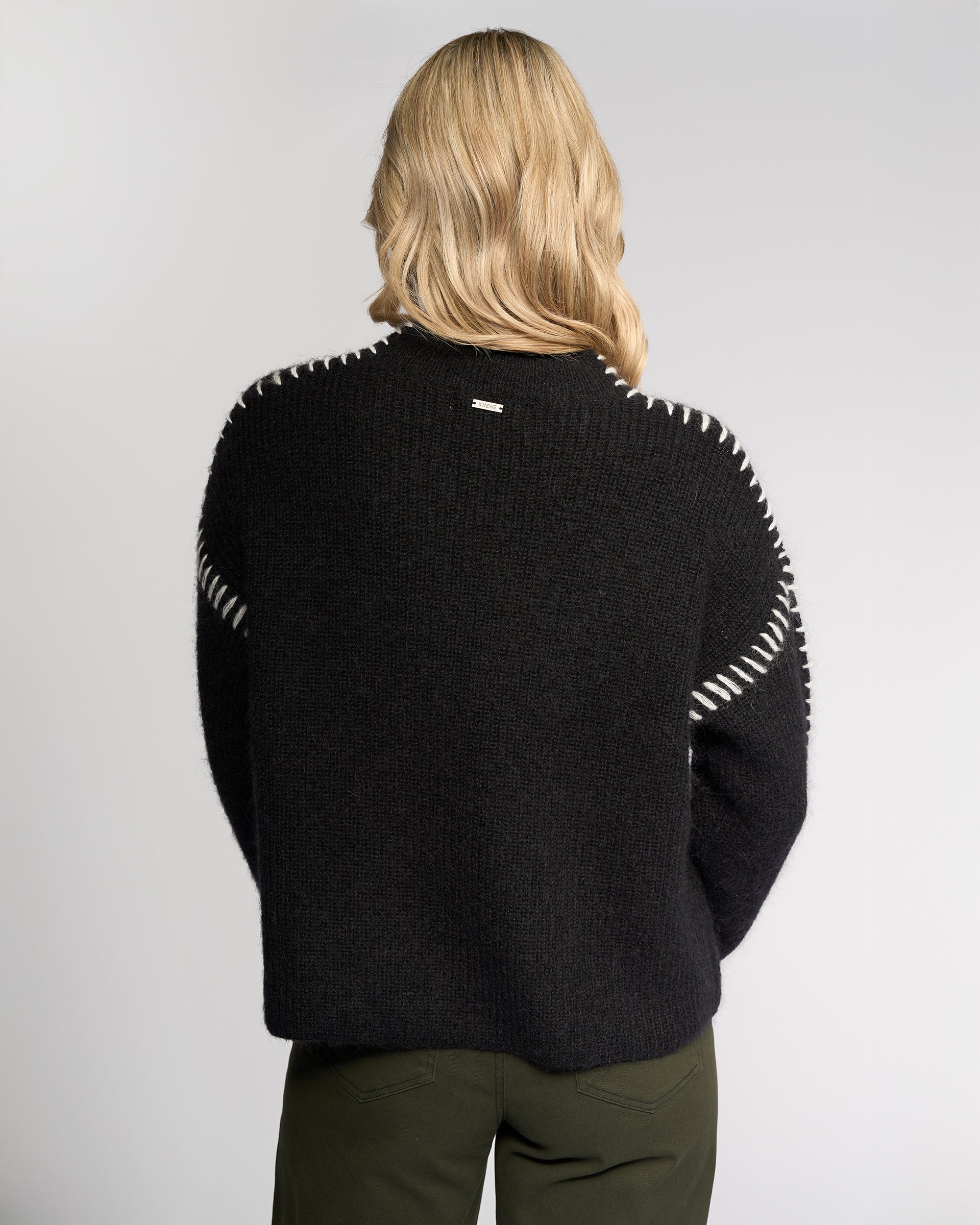 A person with shoulder-length blonde hair is seen from behind wearing the Knewe Label Lennox Sweater in Black/Winter White, featuring relaxed fit and white shoulder stitching, paired with dark green pants against a plain light backdrop.