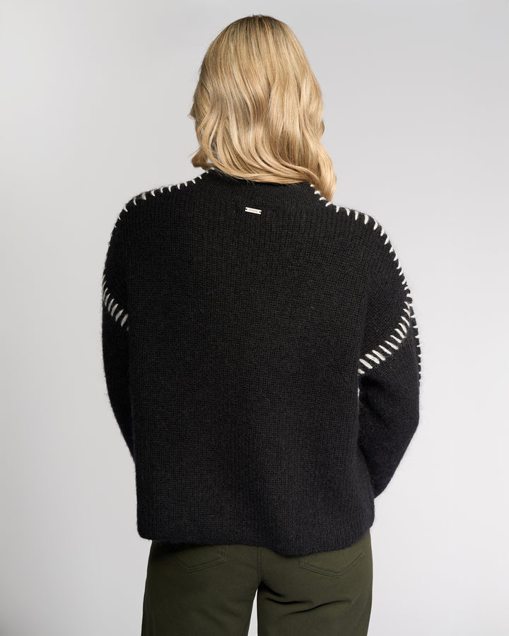 A person with shoulder-length blonde hair is seen from behind wearing the Knewe Label Lennox Sweater in Black/Winter White, featuring relaxed fit and white shoulder stitching, paired with dark green pants against a plain light backdrop.