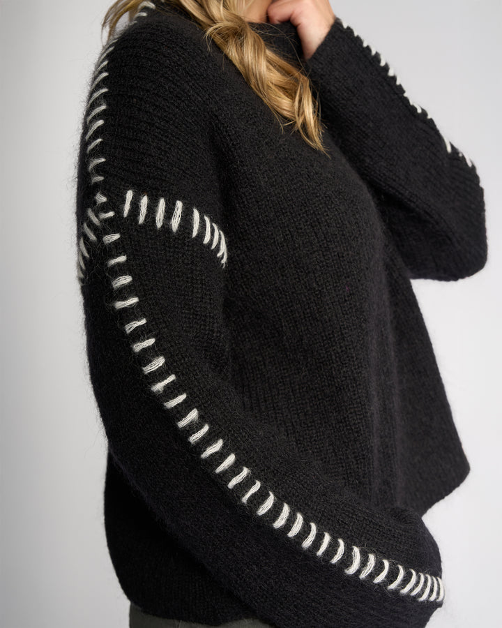 A person wears the Knewe Label Lennox Sweater in Black/Winter White, featuring relaxed fit and thick white stitching along the sleeves and shoulder, shown from the side with their face partially out of frame.