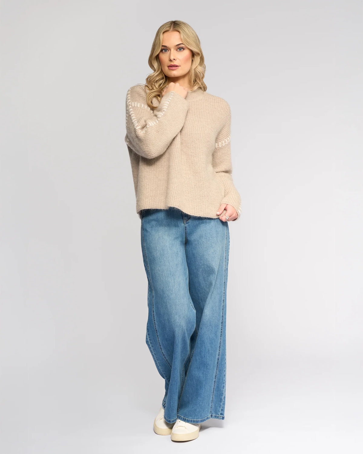 A woman with long blonde hair wears the Lennox Sweater in Oat/Winter White by Knewe Label, paired with wide-leg blue jeans and white sneakers. She stands against a light gray background, touching her hair and looking relaxed.