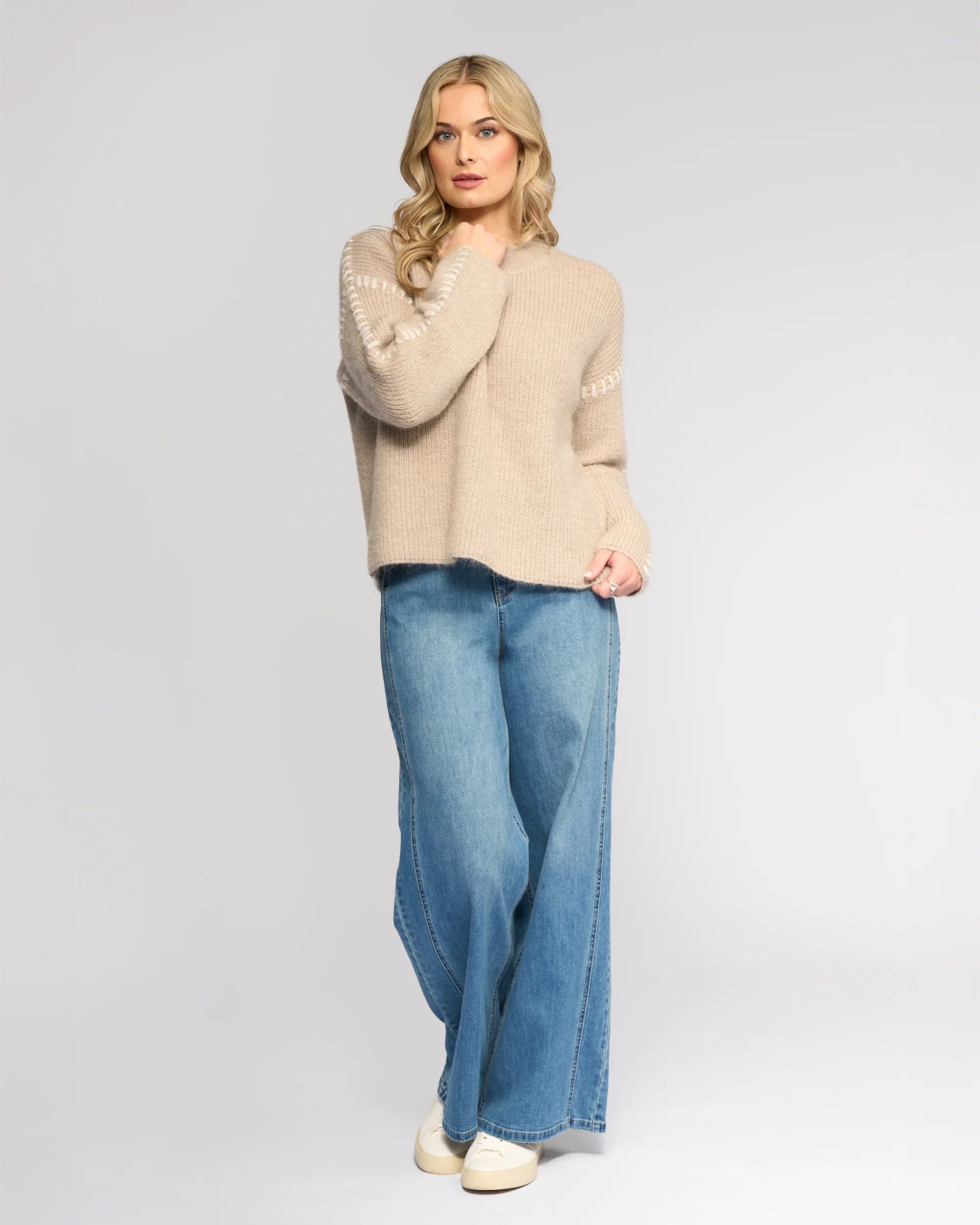 A woman with long blonde hair wears the Lennox Sweater in Oat/Winter White by Knewe Label, paired with wide-leg blue jeans and white sneakers. She stands against a light gray background, touching her hair and looking relaxed.