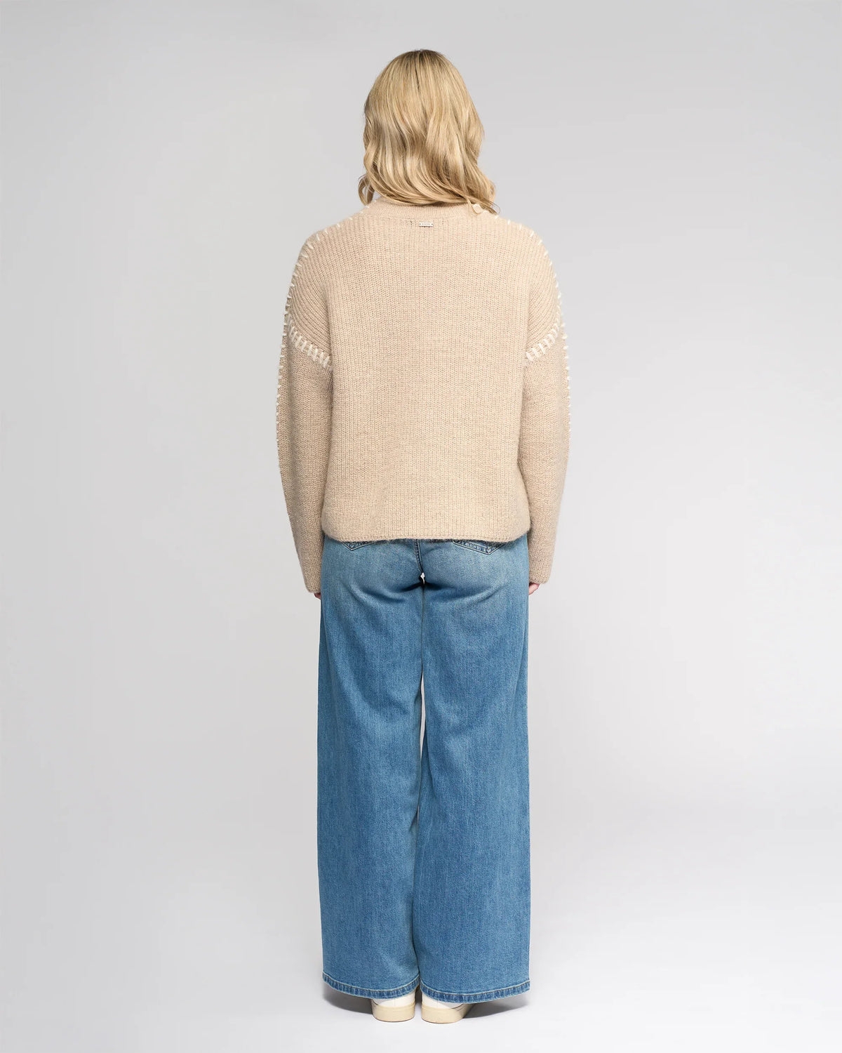 A person with shoulder-length blonde hair stands facing away, wearing the Knewe Label Lennox Sweater in Oat/Winter White, wide-leg blue jeans, and white shoes against a plain light gray background.