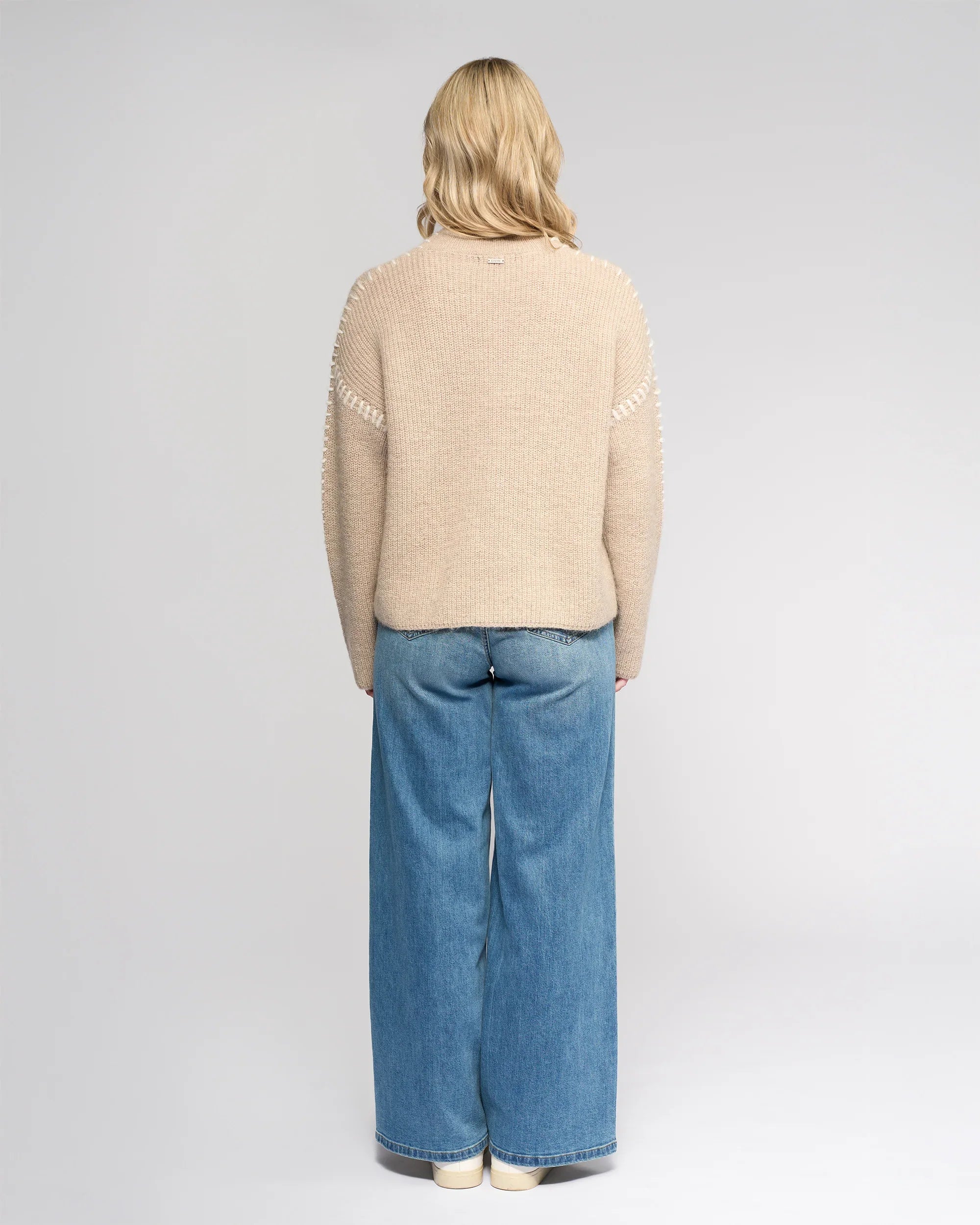 A person with shoulder-length blonde hair stands facing away, wearing the Knewe Label Lennox Sweater in Oat/Winter White, wide-leg blue jeans, and white shoes against a plain light gray background.