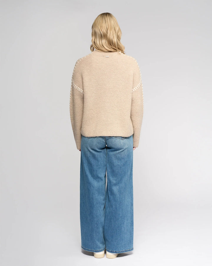 A person with shoulder-length blonde hair stands facing away, wearing the Knewe Label Lennox Sweater in Oat/Winter White, wide-leg blue jeans, and white shoes against a plain light gray background.