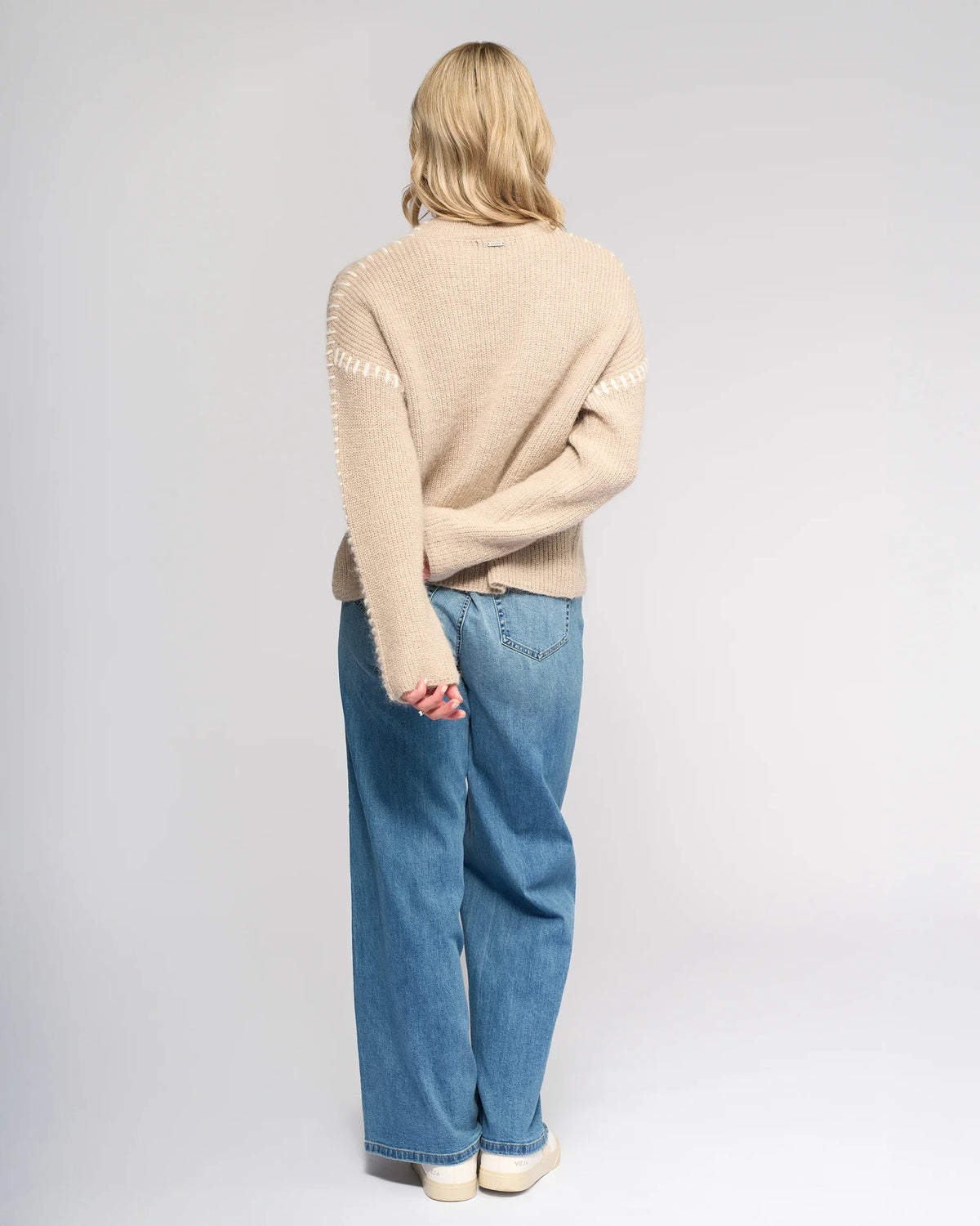 A person with light blond hair wears the Knewe Label Lennox Sweater in Oat/Winter White, paired with loose blue jeans and white shoes, standing facing away from the camera against a plain light background.