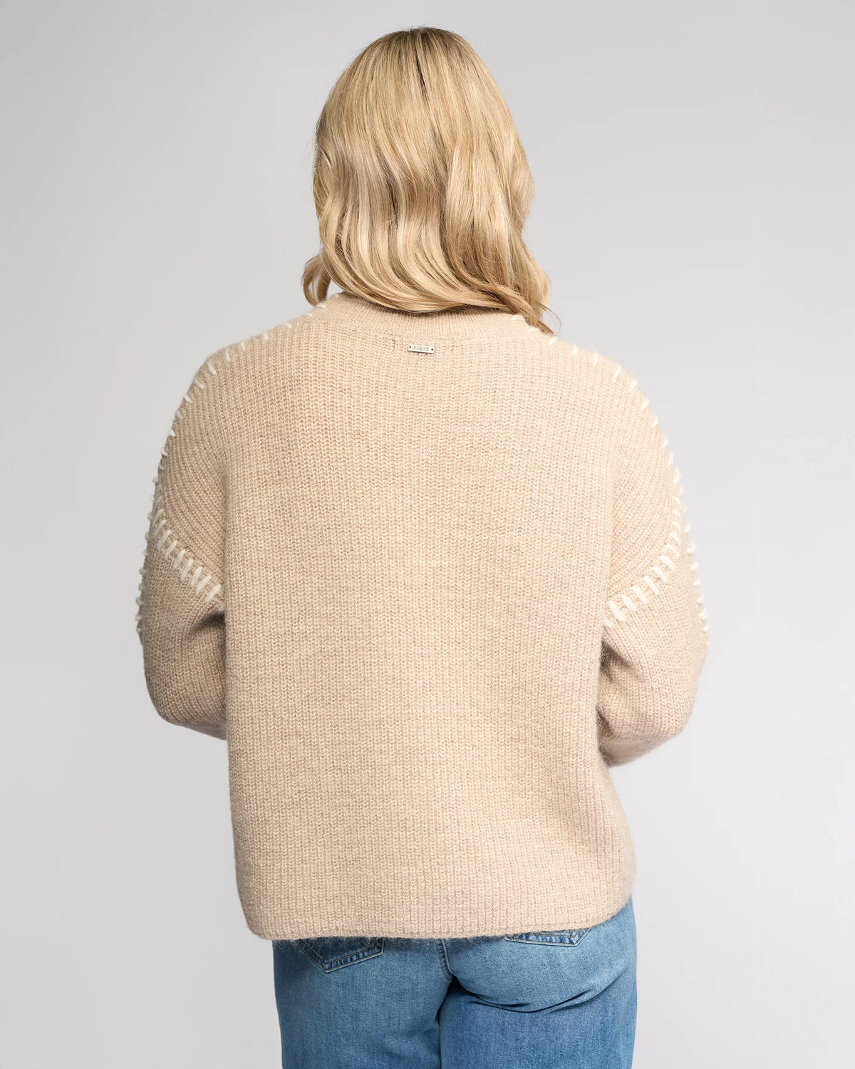 A person with blonde, wavy hair is seen from the back wearing the Knewe Label Lennox Sweater in Oat/Winter White, styled with blue jeans and standing against a light gray backdrop.