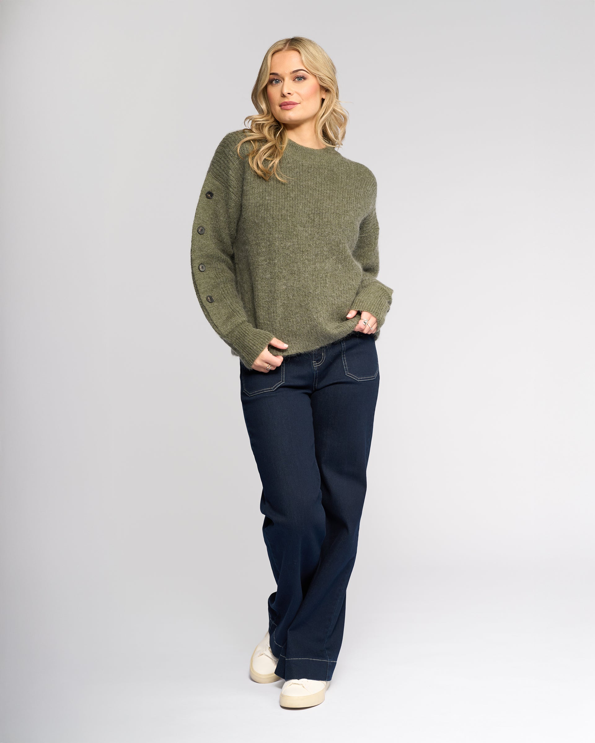 A woman with long blonde hair wears the Knewe Label Luca Sweater in Elm, paired with dark blue jeans and white sneakers. She stands against a plain light background, one hand in her pocket, looking at the camera.
