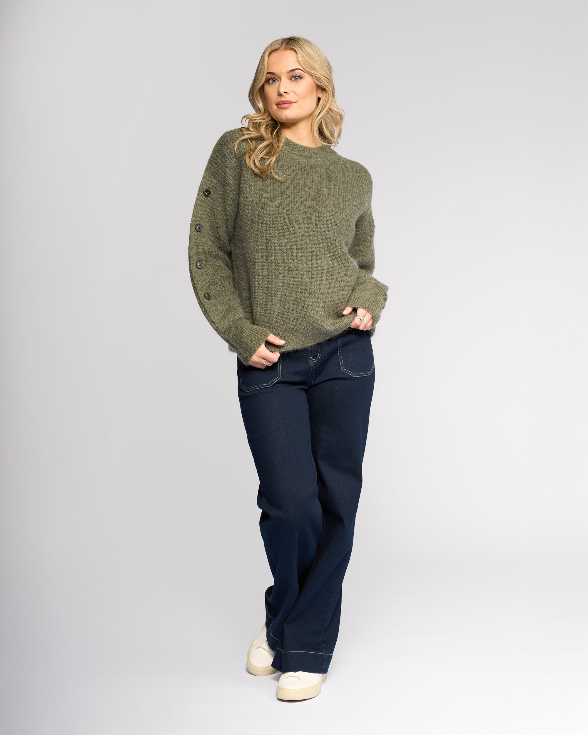 A woman with long blonde hair wears the Knewe Label Luca Sweater in Elm, paired with dark blue jeans and white sneakers. She stands against a plain light background, one hand in her pocket, looking at the camera.