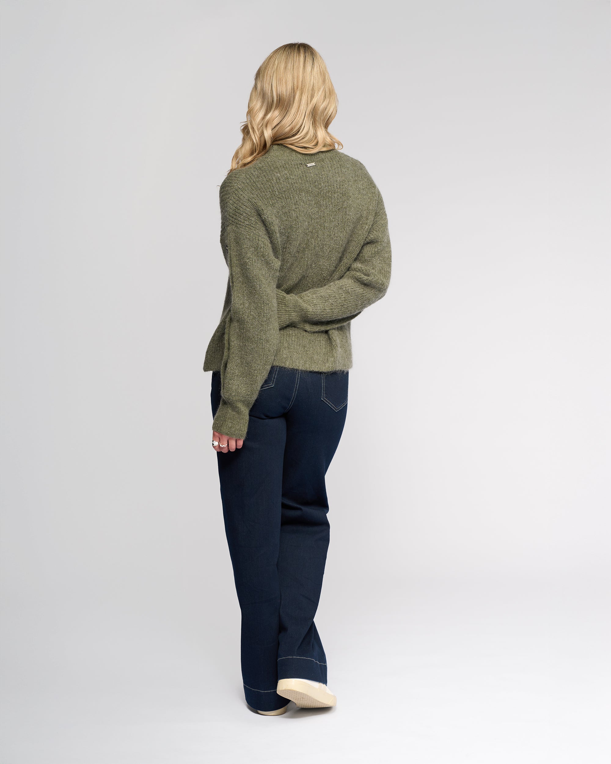 A person with long blonde hair, wearing the Knewe Label Luca Sweater in Elm and dark jeans, stands facing away from the camera against a plain light background.
