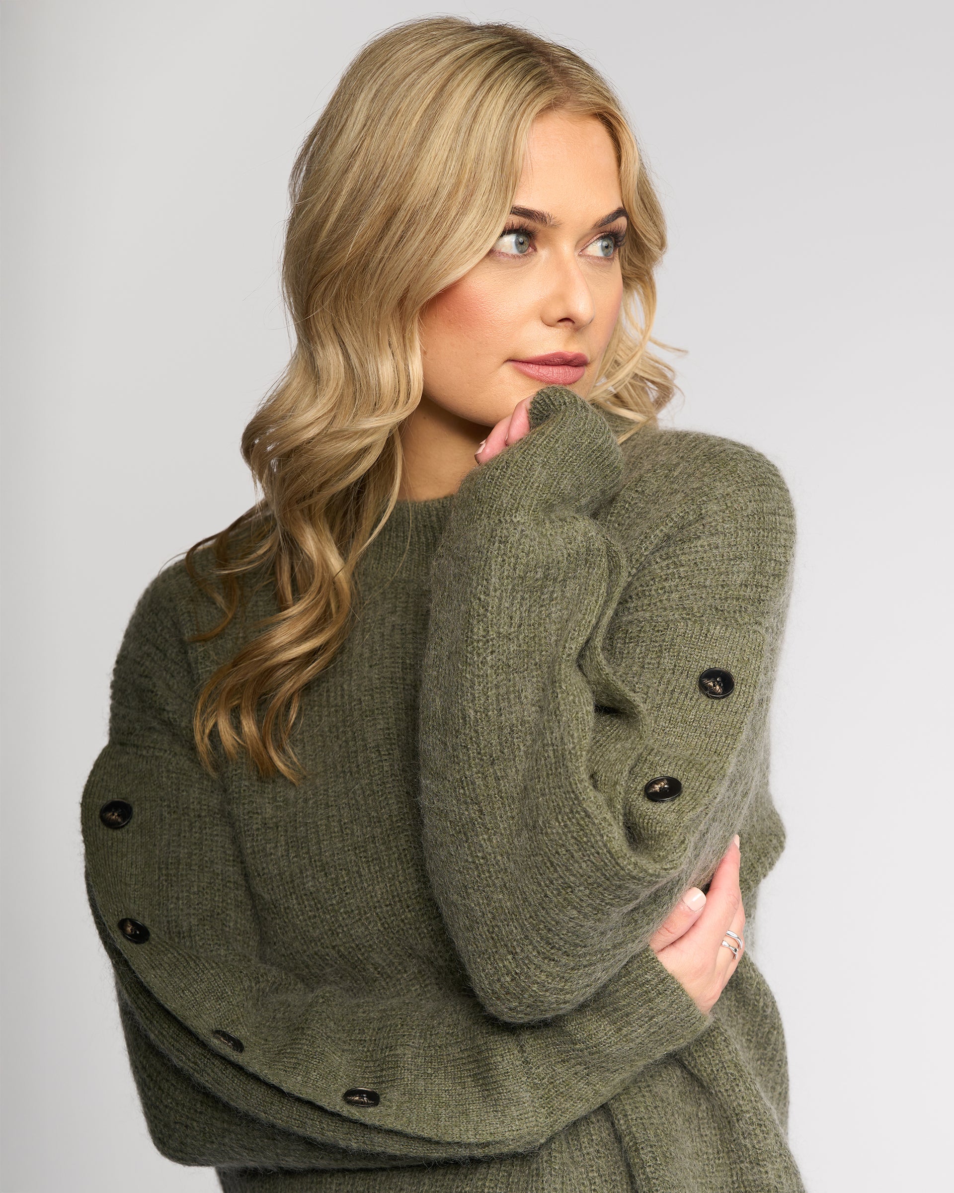 A woman with long, wavy blonde hair wears the Luca Sweater - Elm by Knewe Label, featuring cozy mohair wool and black buttoned split sleeves, posing thoughtfully with arms crossed against a plain light background.