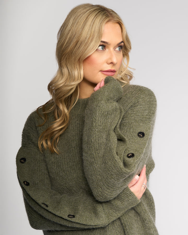 A woman with long, wavy blonde hair wears the Luca Sweater - Elm by Knewe Label, featuring cozy mohair wool and black buttoned split sleeves, posing thoughtfully with arms crossed against a plain light background.