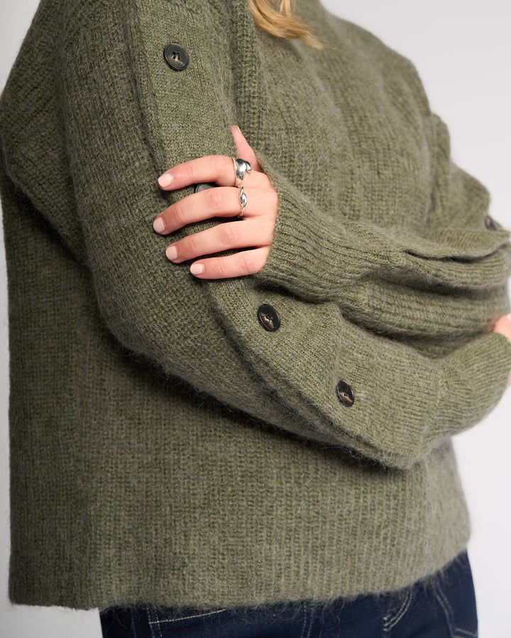 A person wearing the Knewe Label Luca Sweater in Elm, featuring buttoned split sleeves and a textured mohair wool knit, crosses their arms. Silver rings and dark blue jeans complete the look; only the torso and arms are visible.
