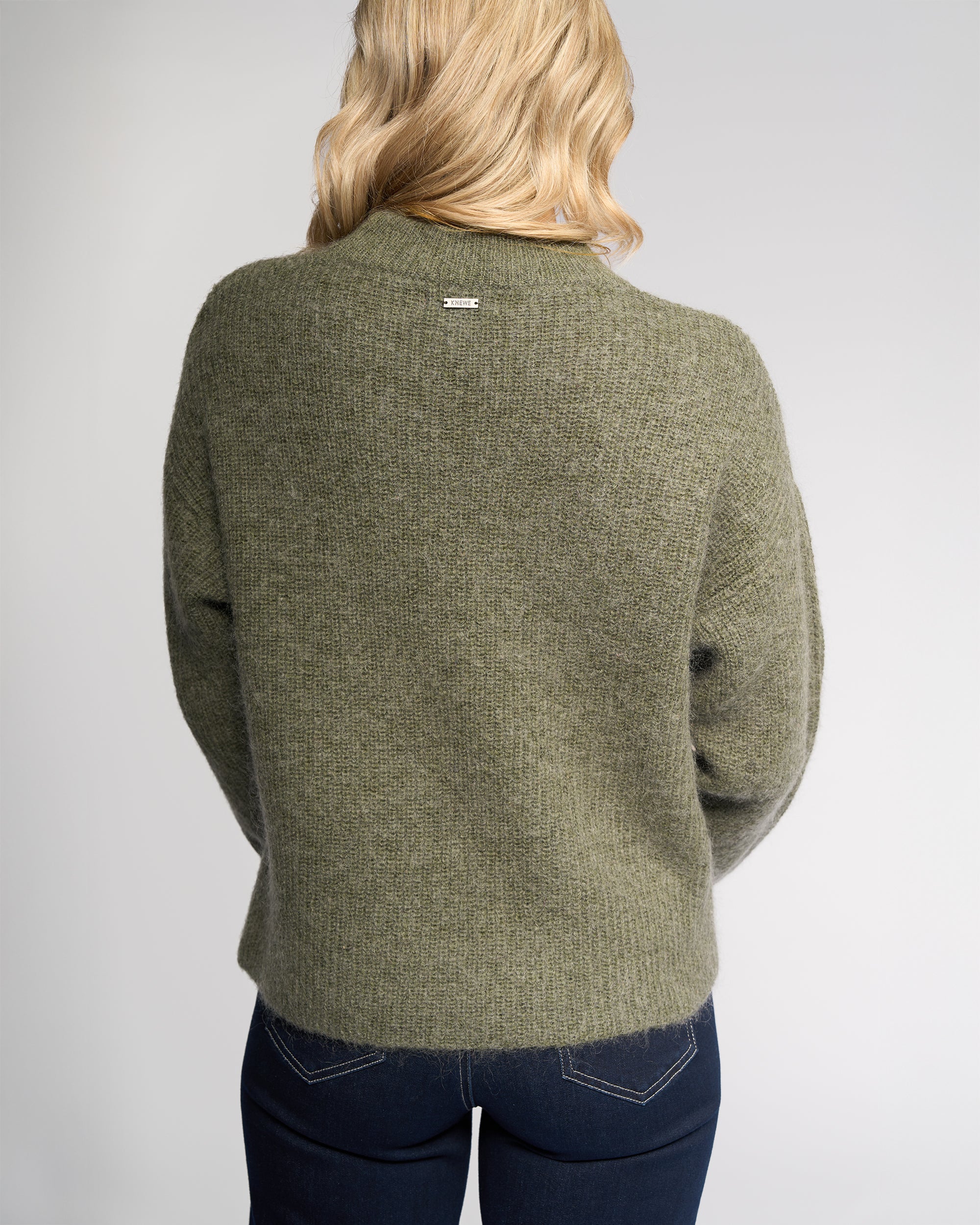 A person with wavy blonde hair is seen from the back, wearing the Luca Sweater in Elm by Knewe Label and dark blue jeans, set against a plain light background.