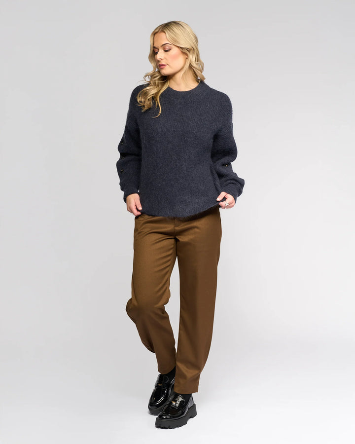 A woman with long blonde hair wears the Luca Sweater - Ink by Knewe Label, paired with brown trousers and black shoes. She stands against a plain light gray background, looking down to her left with one hand in her pocket.
