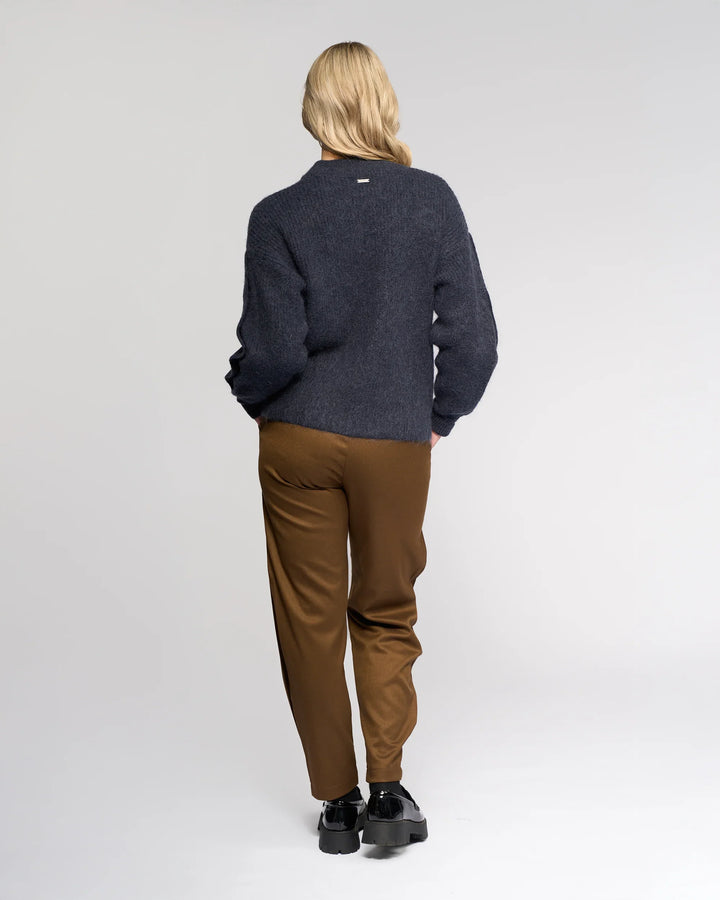 A person with long blonde hair faces away, wearing the Knewe Label Luca Sweater - Ink with buttoned split sleeves, brown pants, and black shoes, standing against a plain light gray background.