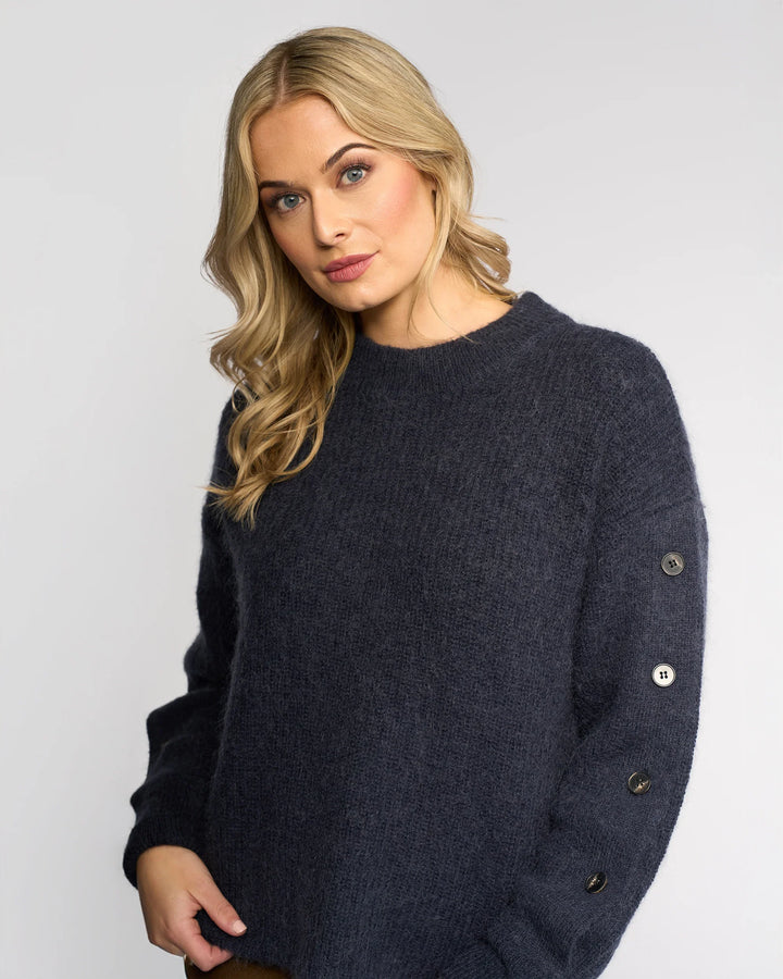 A woman with long blonde hair wears the Knewe Label Luca Sweater in Ink, featuring buttoned split sleeves, against a plain light background, looking slightly to the side with a neutral expression.