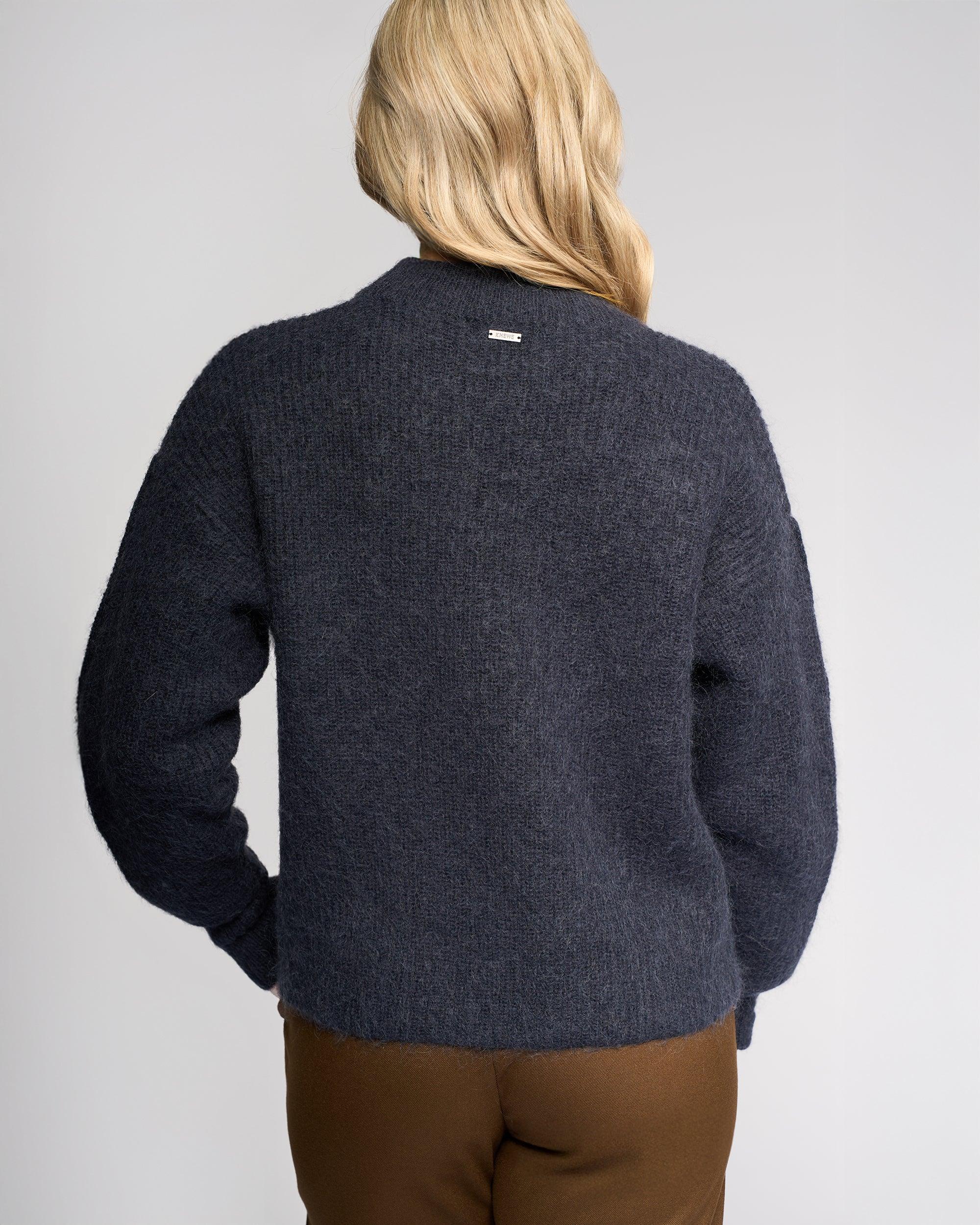 A person with long blonde hair faces away from the camera, wearing Knewe Label’s Luca Sweater in Ink—a dark navy mohair wool sweater with buttoned split sleeves—paired with brown pants against a plain light gray background.