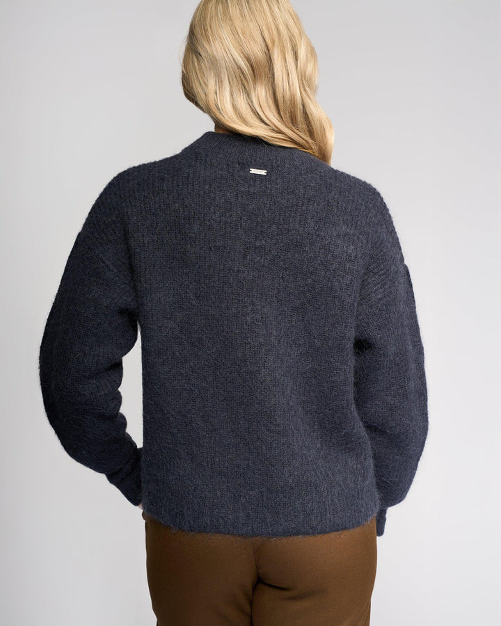 A person with long blonde hair faces away from the camera, wearing Knewe Label’s Luca Sweater in Ink—a dark navy mohair wool sweater with buttoned split sleeves—paired with brown pants against a plain light gray background.