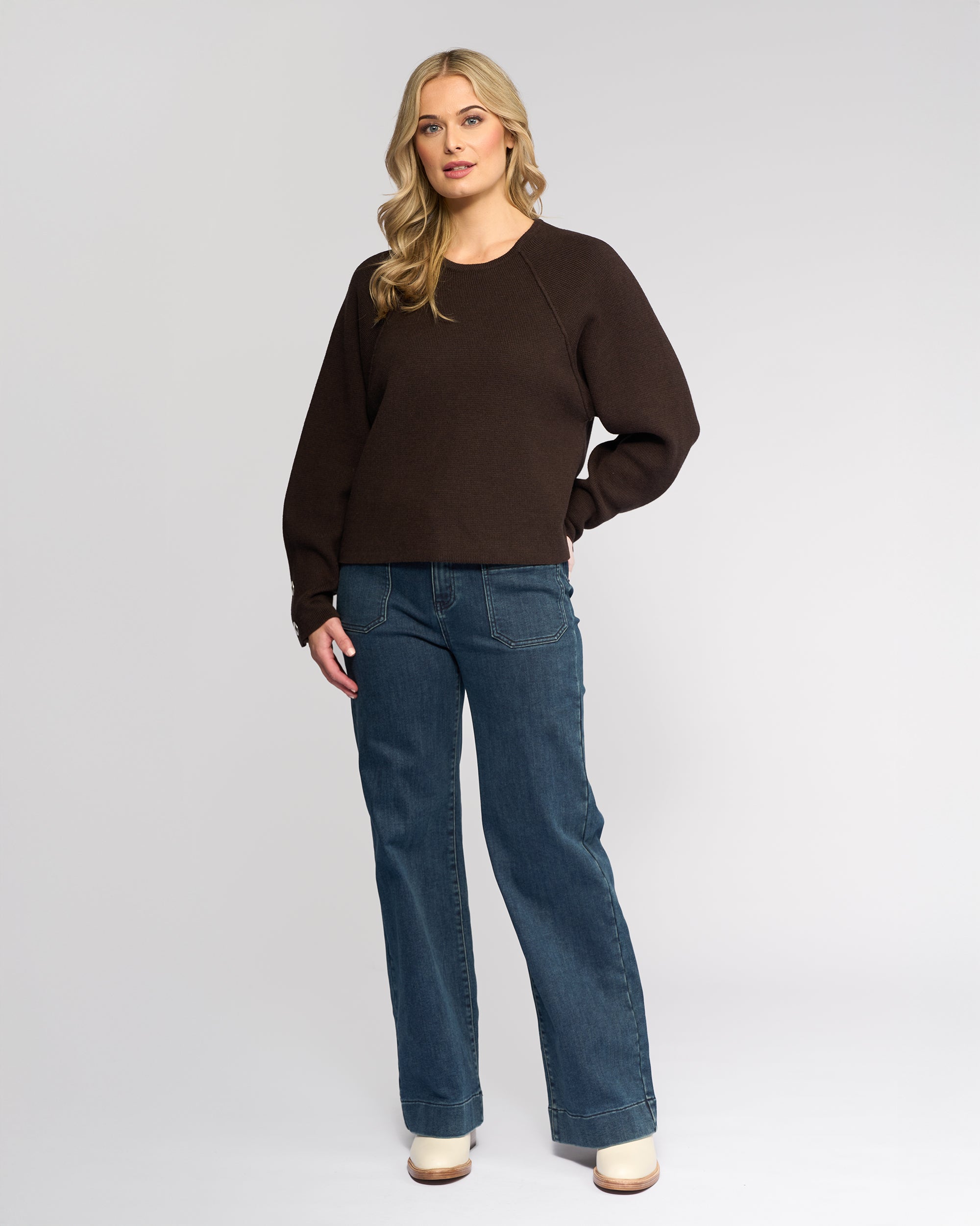 A woman with long blonde hair wears the Knewe Label Juno Sweater in Chocolate, paired with blue jeans and cream-colored shoes, standing against a plain light gray background.