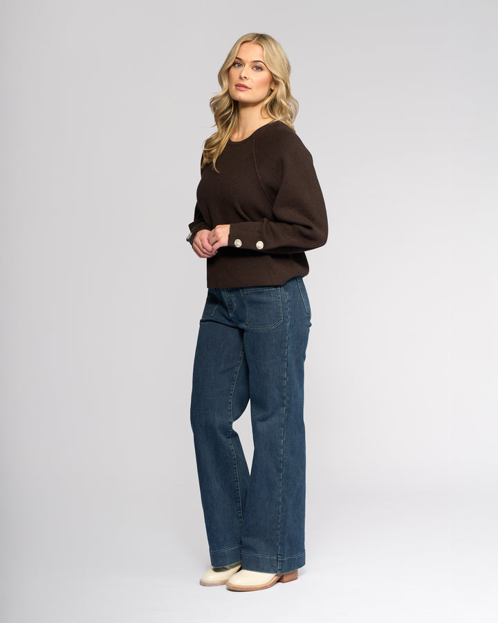 A woman with long blonde hair stands against a light background, wearing the Knewe Label Juno Sweater in Chocolate with oversized sleeves and cuff buttons, styled with blue wide-leg jeans and cream shoes as she looks toward the camera.