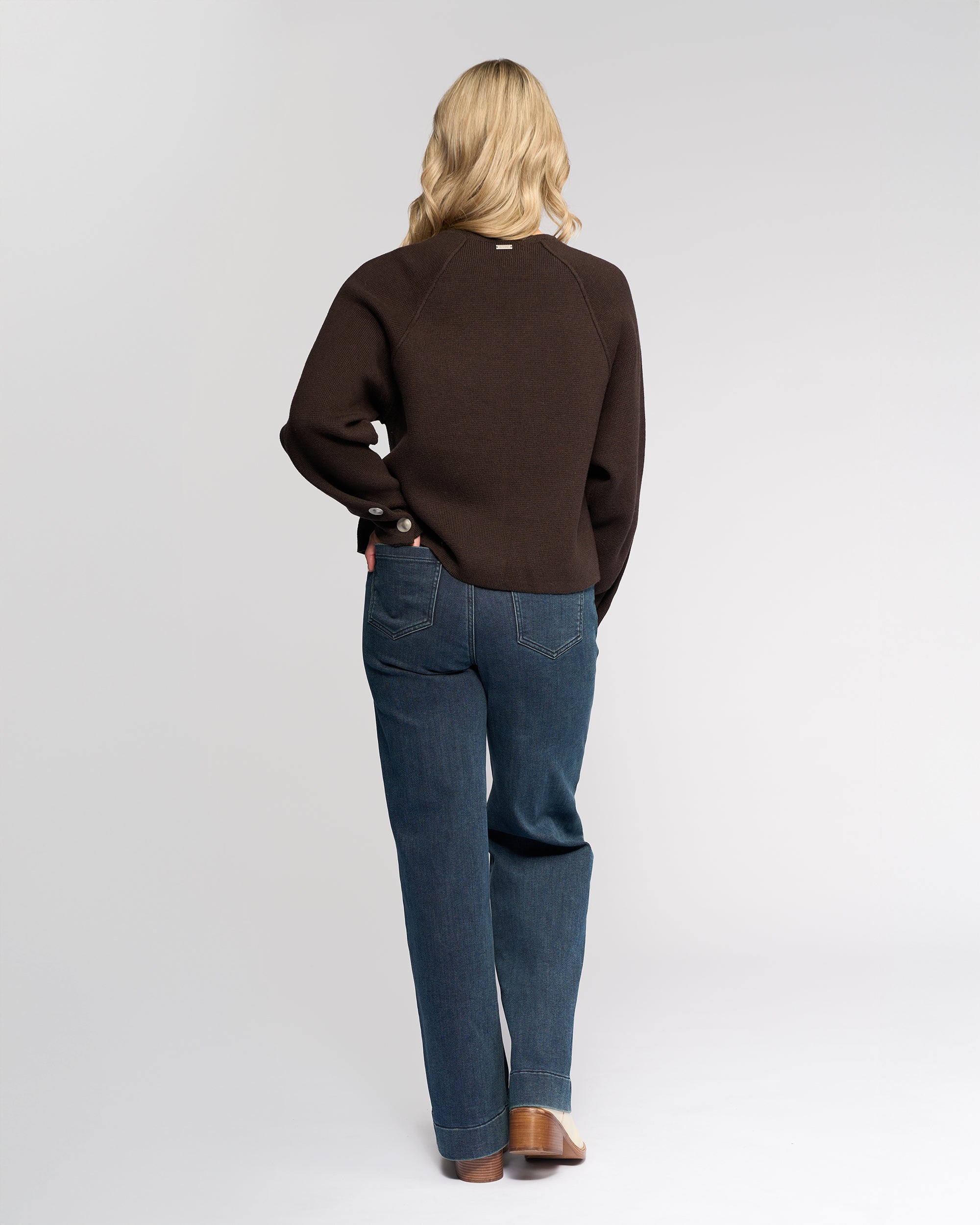 A person with long blonde hair, wearing the Knewe Label Juno Sweater in Chocolate, blue jeans, and brown shoes, stands facing away from the camera against a plain light background.